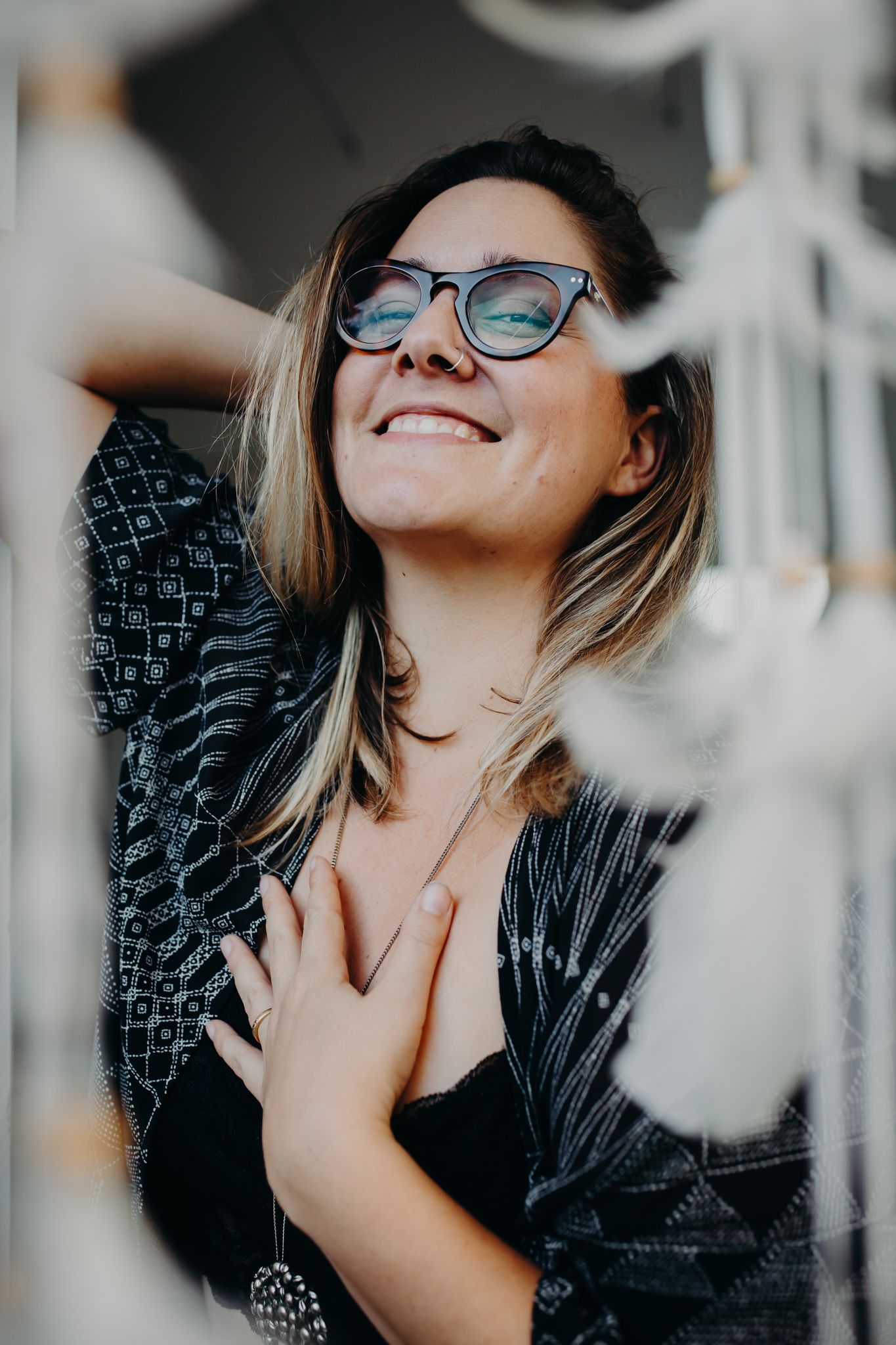 Candid portrait of person wearing blue glasses and black top smiling joyfully against blurred background.