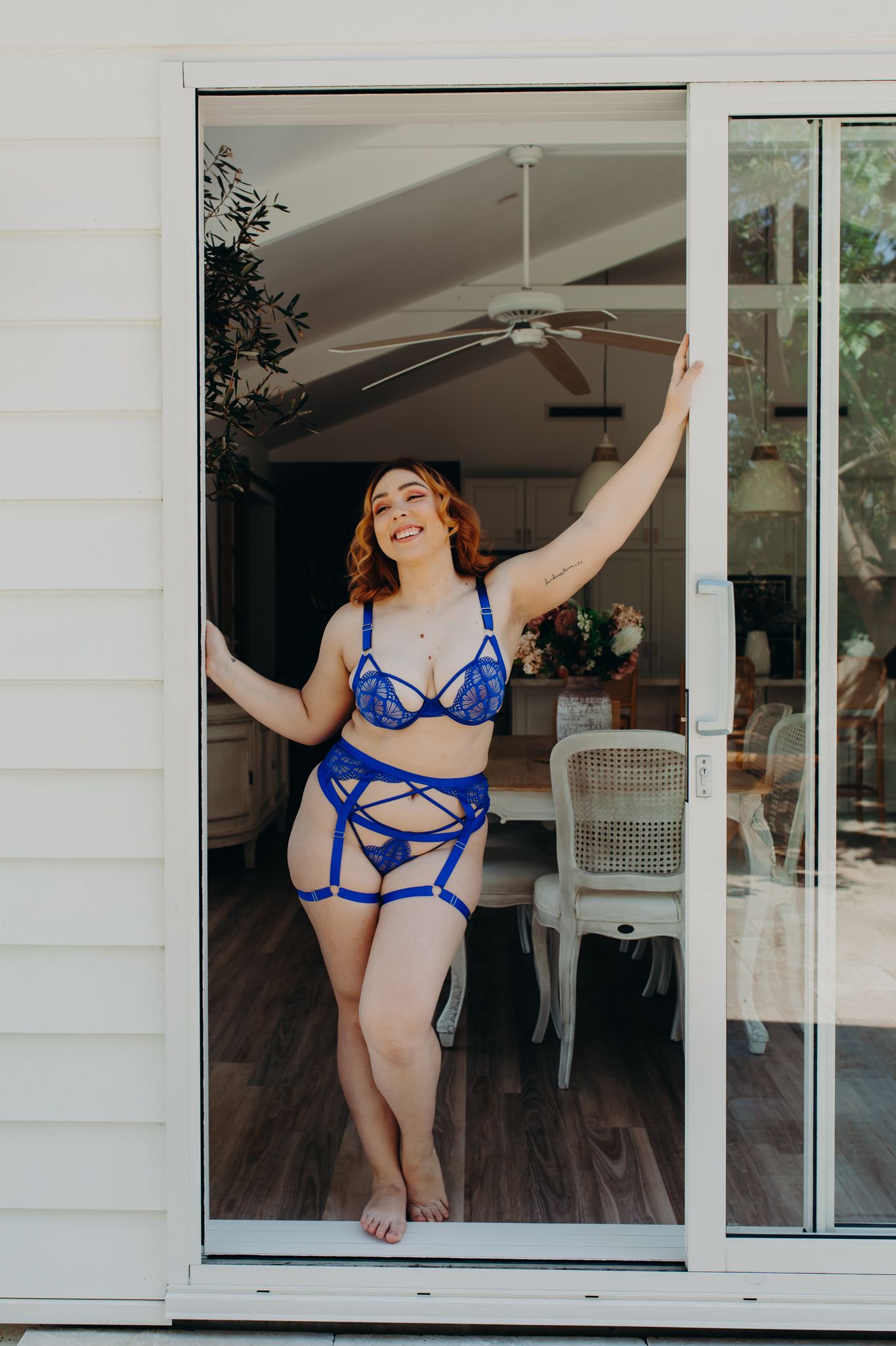 Joyful pose in blue strappy lingerie set by sliding glass door.