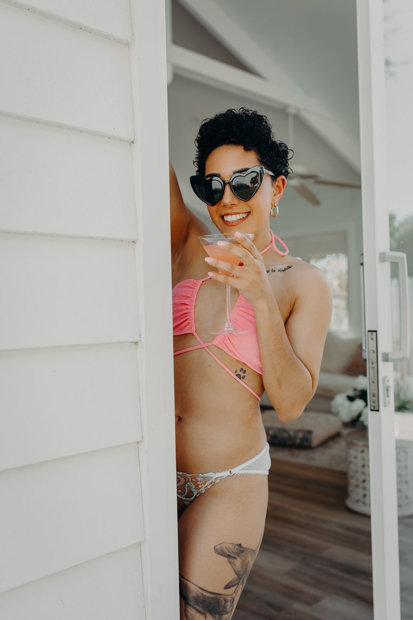 Model in pink bikini and sunglasses poses with drink by white wall.