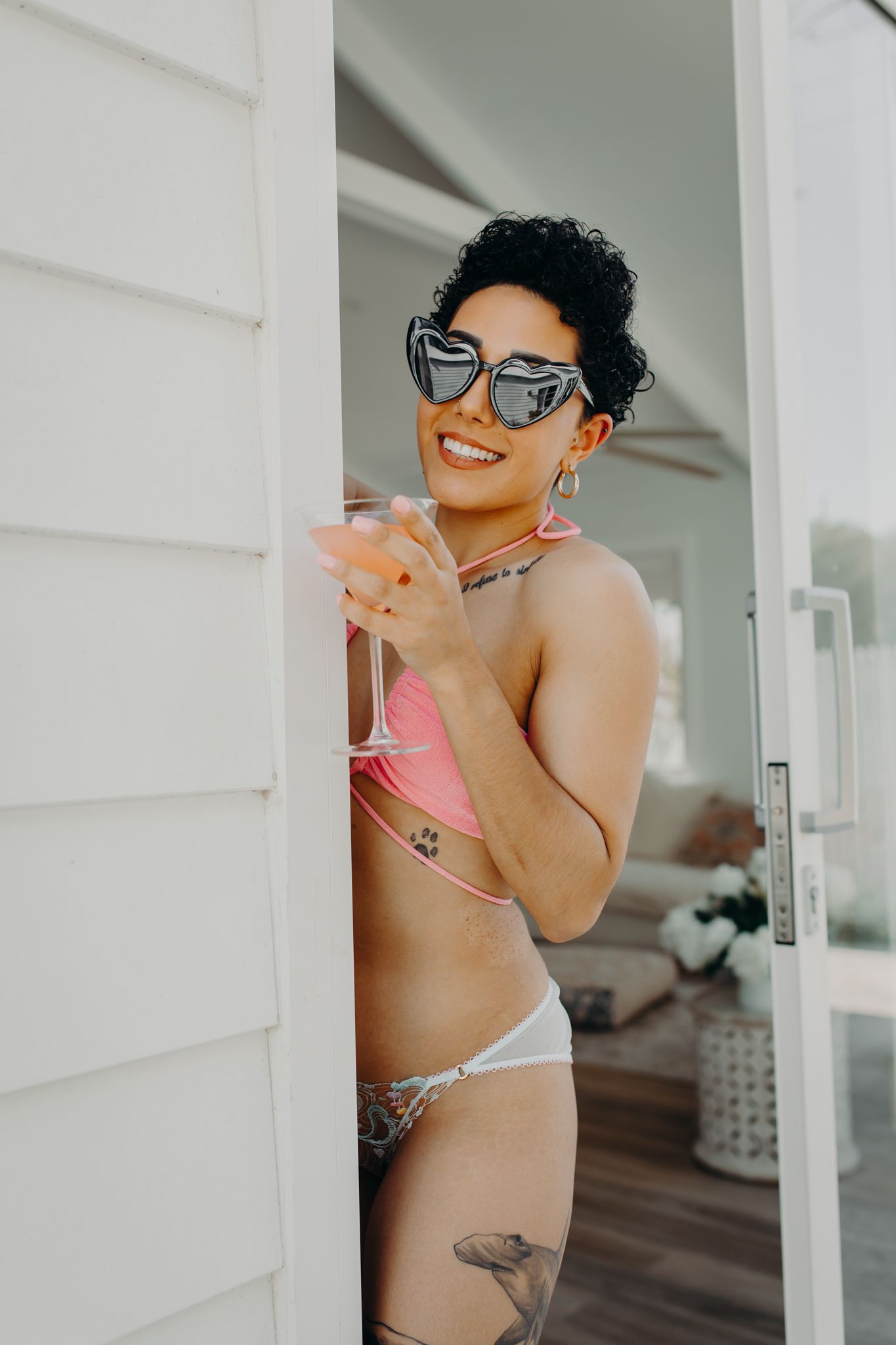 Laughing model in sunglasses and pink swimwear peeks from behind white wall.