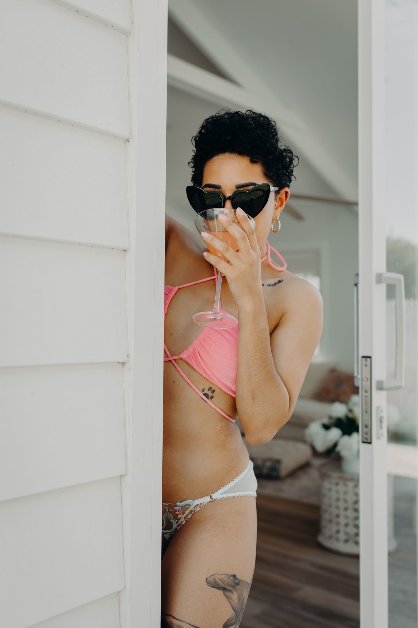Playful model in swimwear and sunglasses poses behind white wall outdoors.