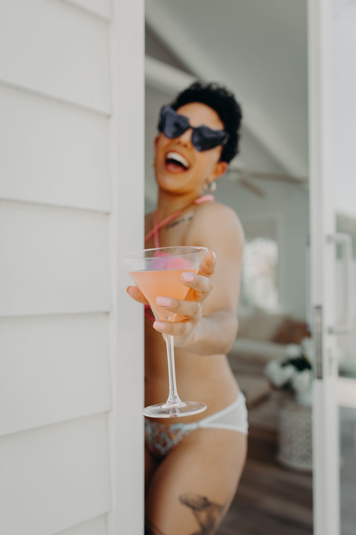 Model holding martini glass poses in swimwear and sunglasses by white exterior wall.