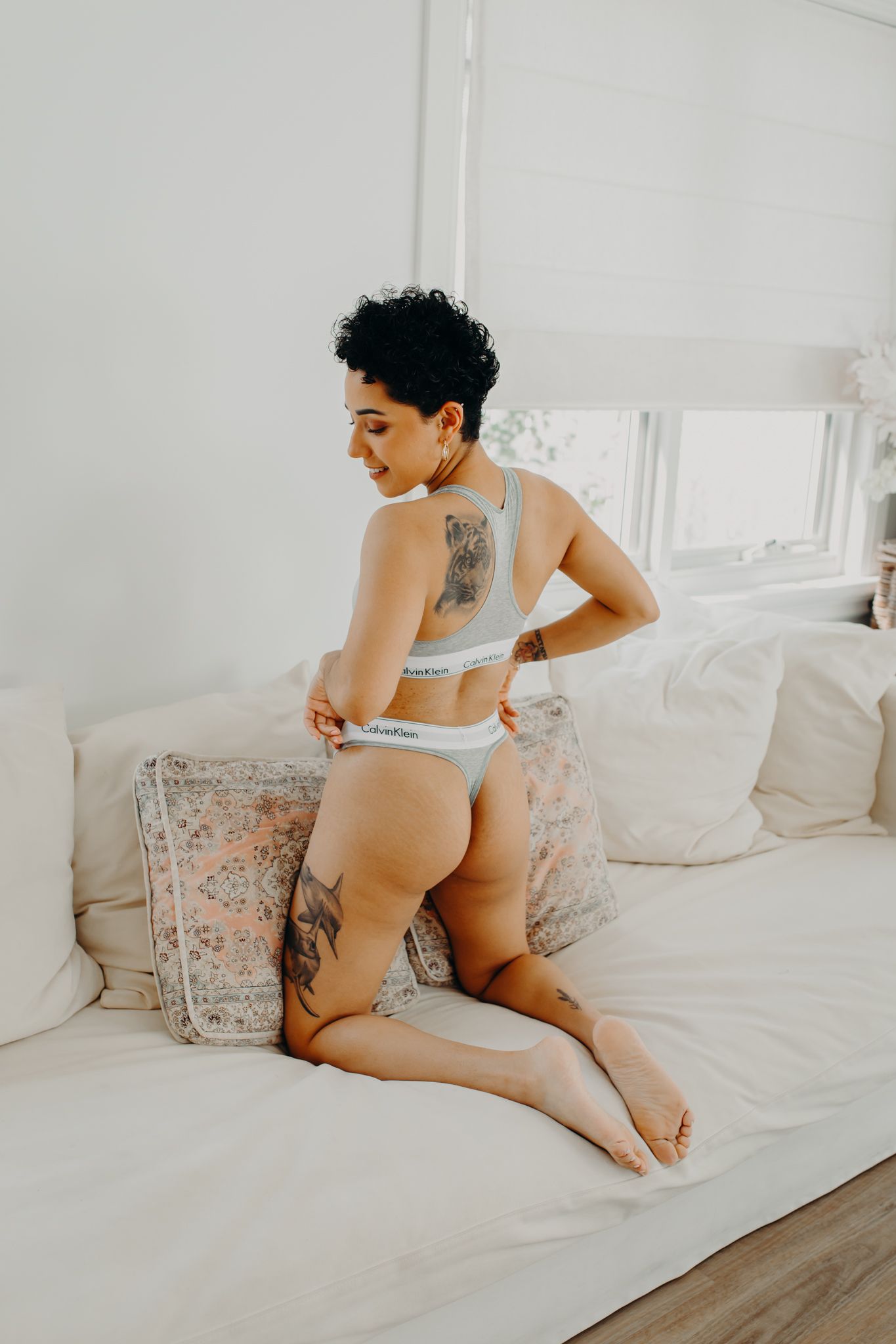 Model wearing Calvin Klein lingerie poses on white sofa showing back tattoo.