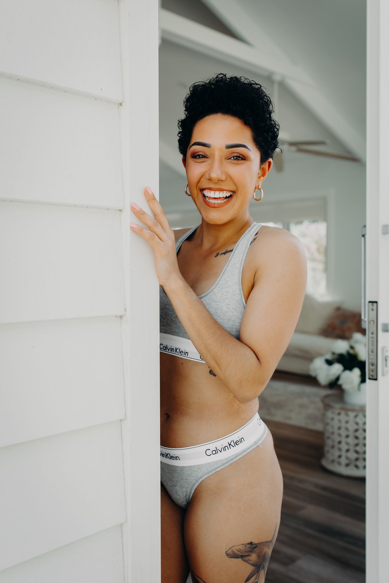 Joyful model in grey Calvin Klein set poses playfully by white doorframe.