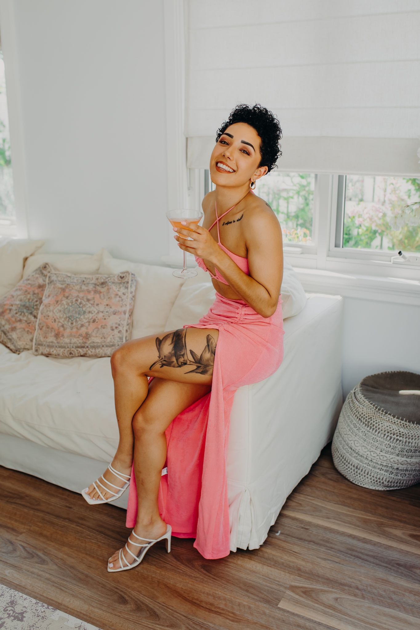 Model in pink dress poses on white couch with cocktail glass in bright interior setting.