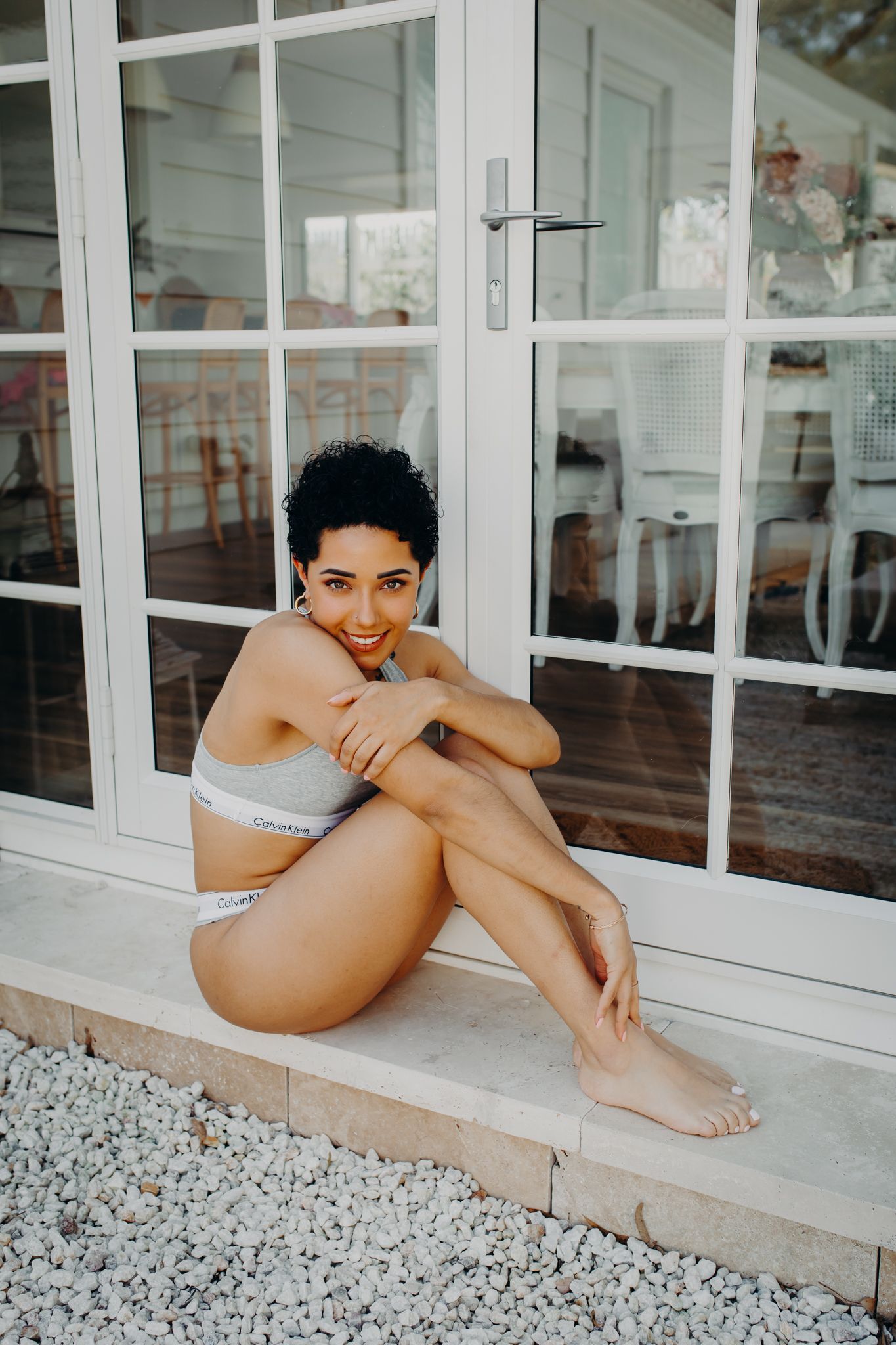 Model in grey underwear sits casually on doorstep against glass doors.