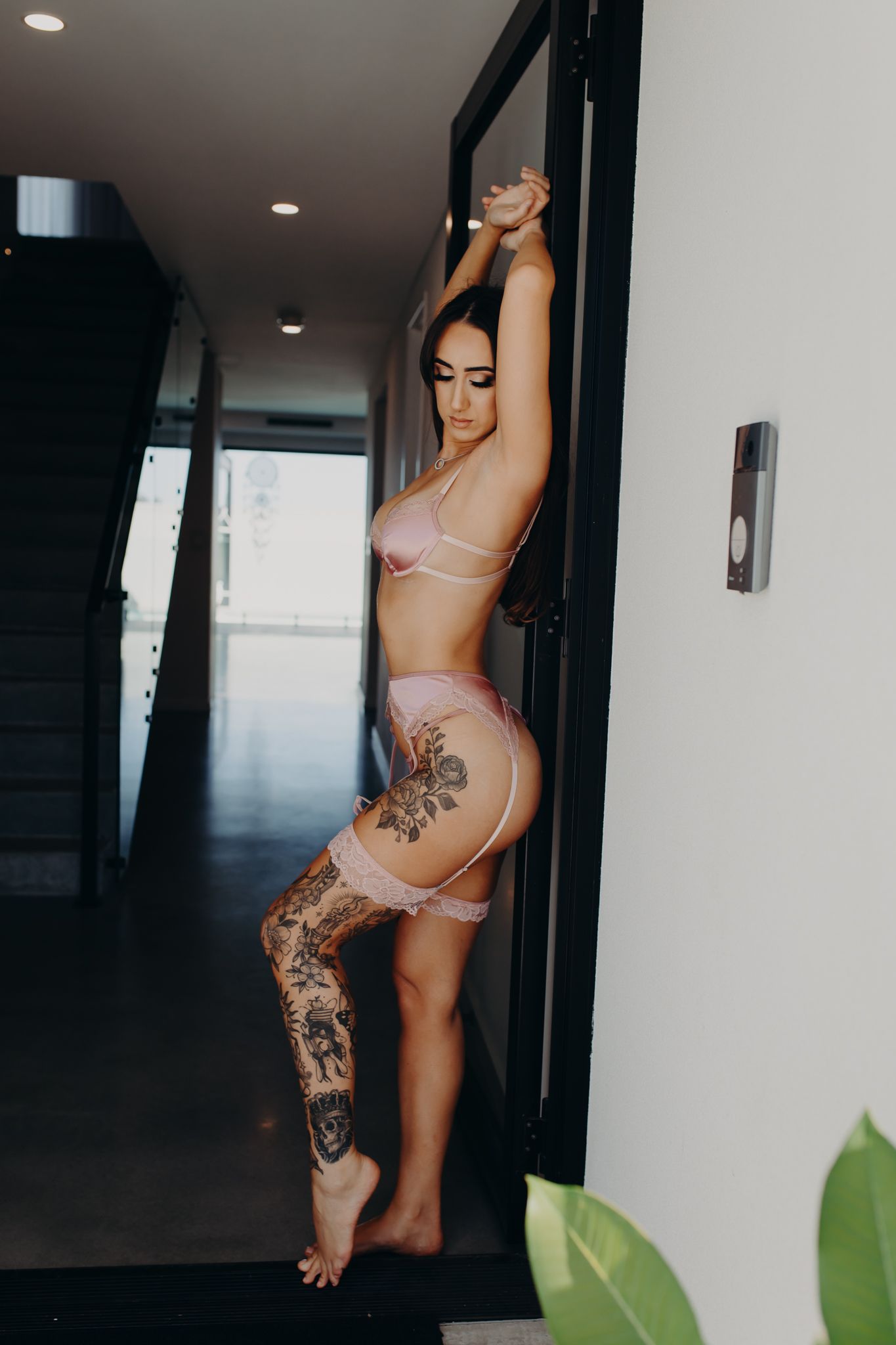 Person in pink lingerie set showing artistic leg tattoos while posing in modern hallway.