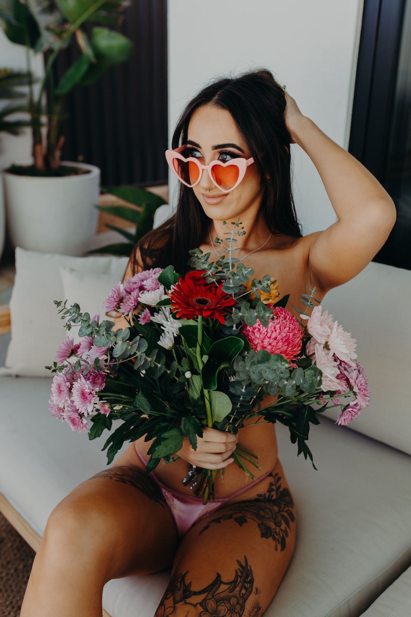 Portrait with heart-shaped sunglasses and colorful flower bouquet on white couch.