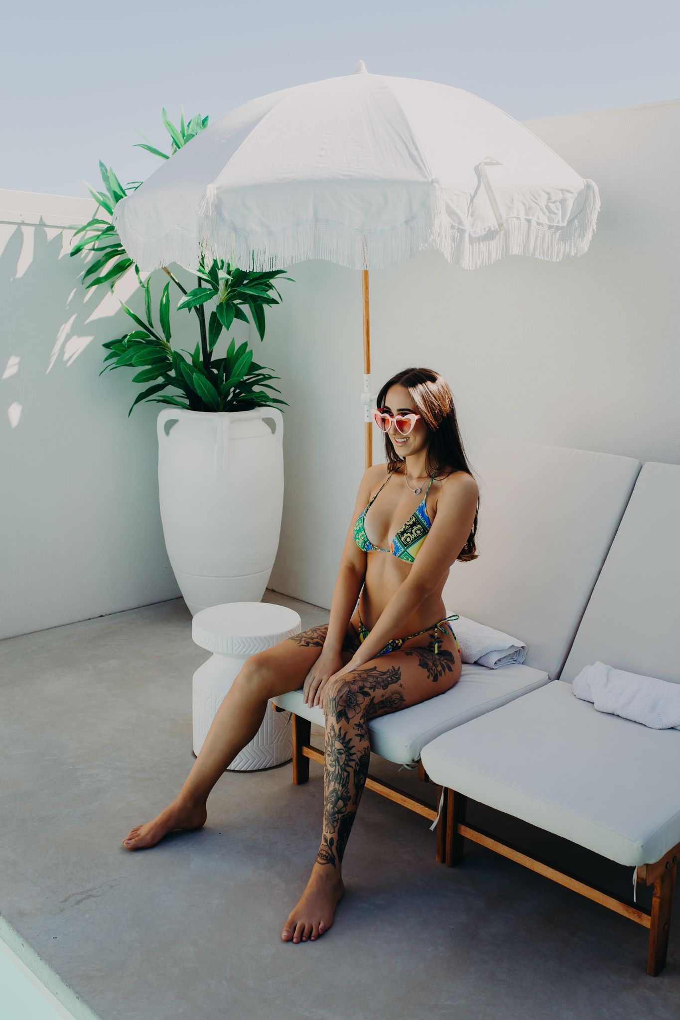 Relaxing poolside scene with bikini and white umbrella next to potted tropical plant on concrete patio.