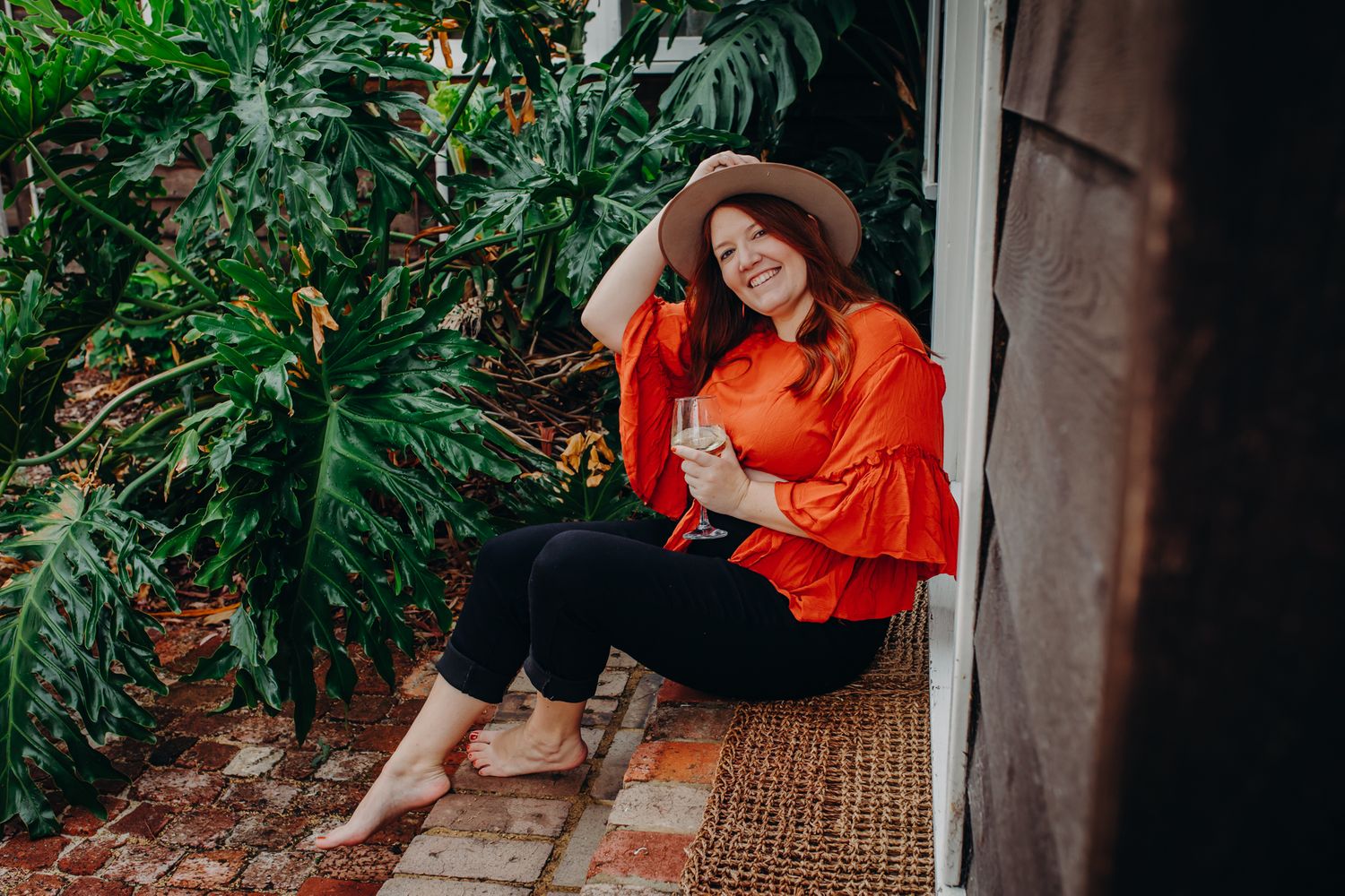 Woman in orange top and brown hat poses on doorstep next to tropical plants and greenery.