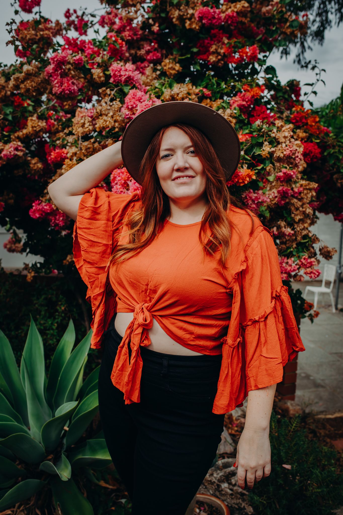 Woman wearing orange ruffled top and brown hat poses in front of blooming pink bougainvillea flowers.