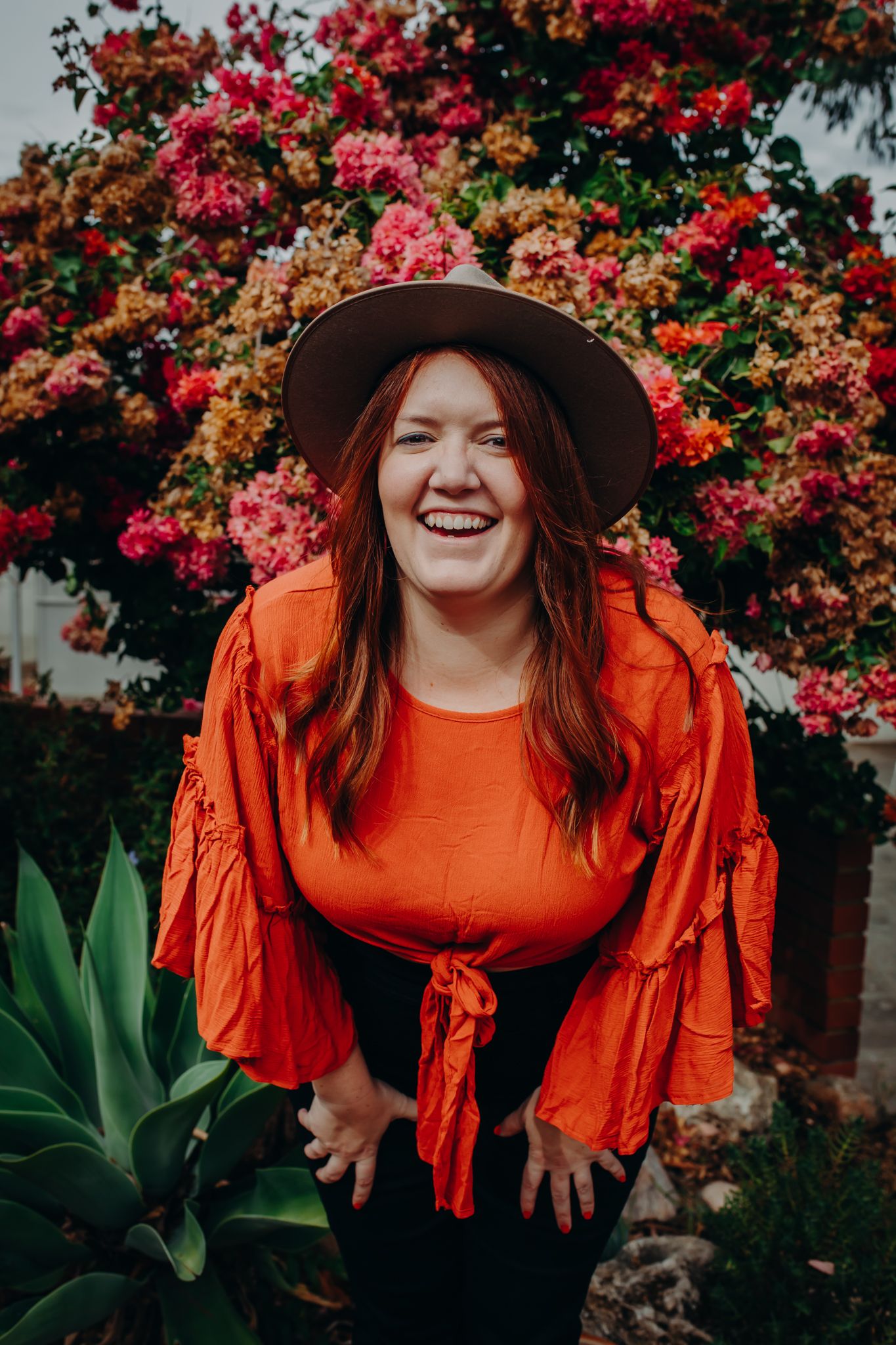 Cheerful woman with brown sun hat and orange top enjoys a sunny moment among blooming flowers.