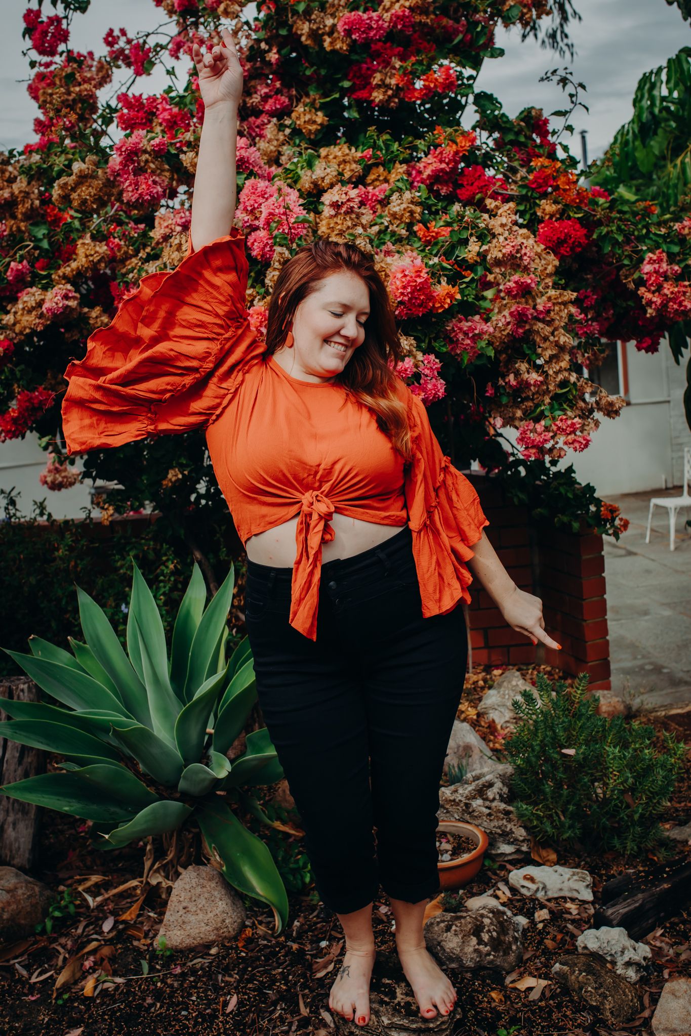 Woman in orange crop top raises arm joyfully against backdrop of pink flowers and succulents.