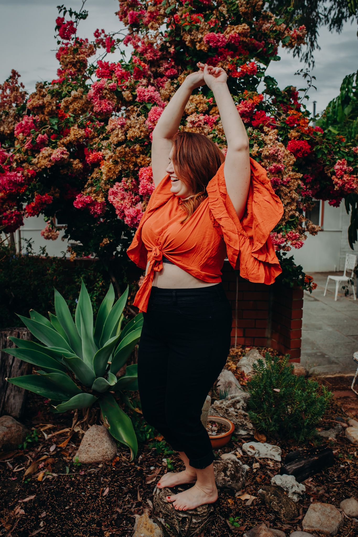 Woman in orange ruffled top stretches against garden backdrop with bougainvillea and agave plants.