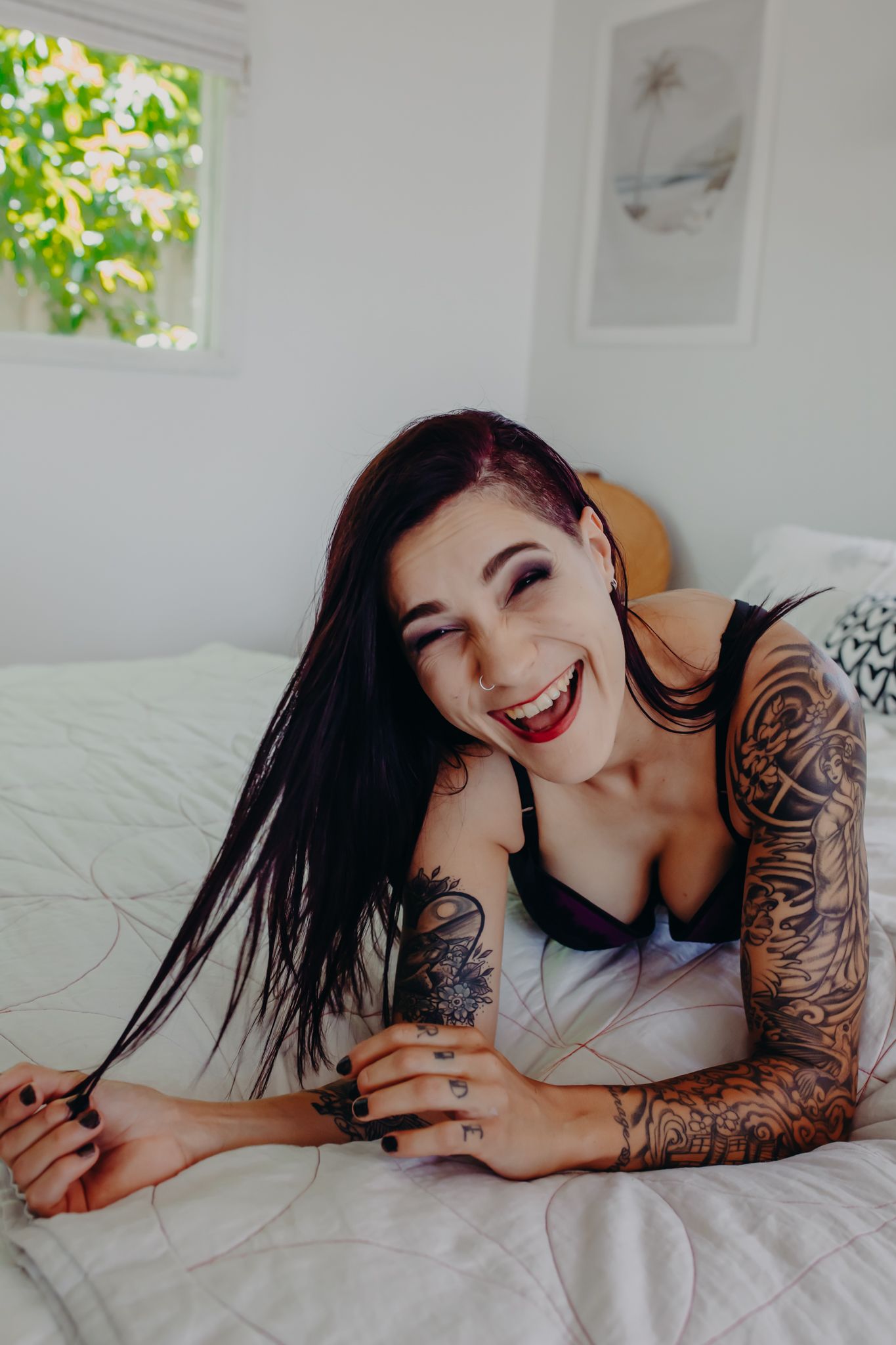 Model with sleeve tattoo laughs while lying on white bedding in casual boudoir pose.
