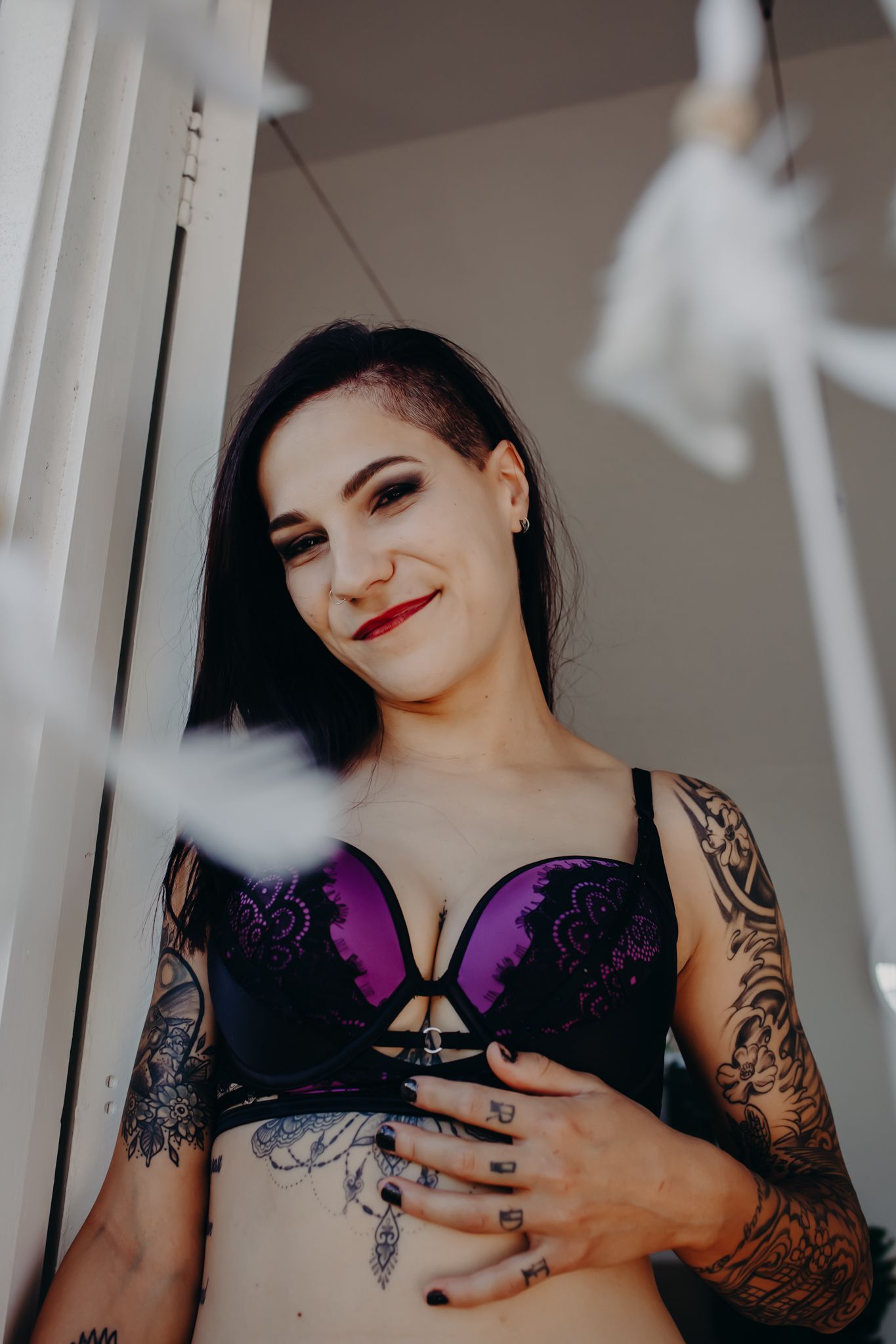 Model in purple lingerie poses by window with visible sleeve tattoo and dark hair.