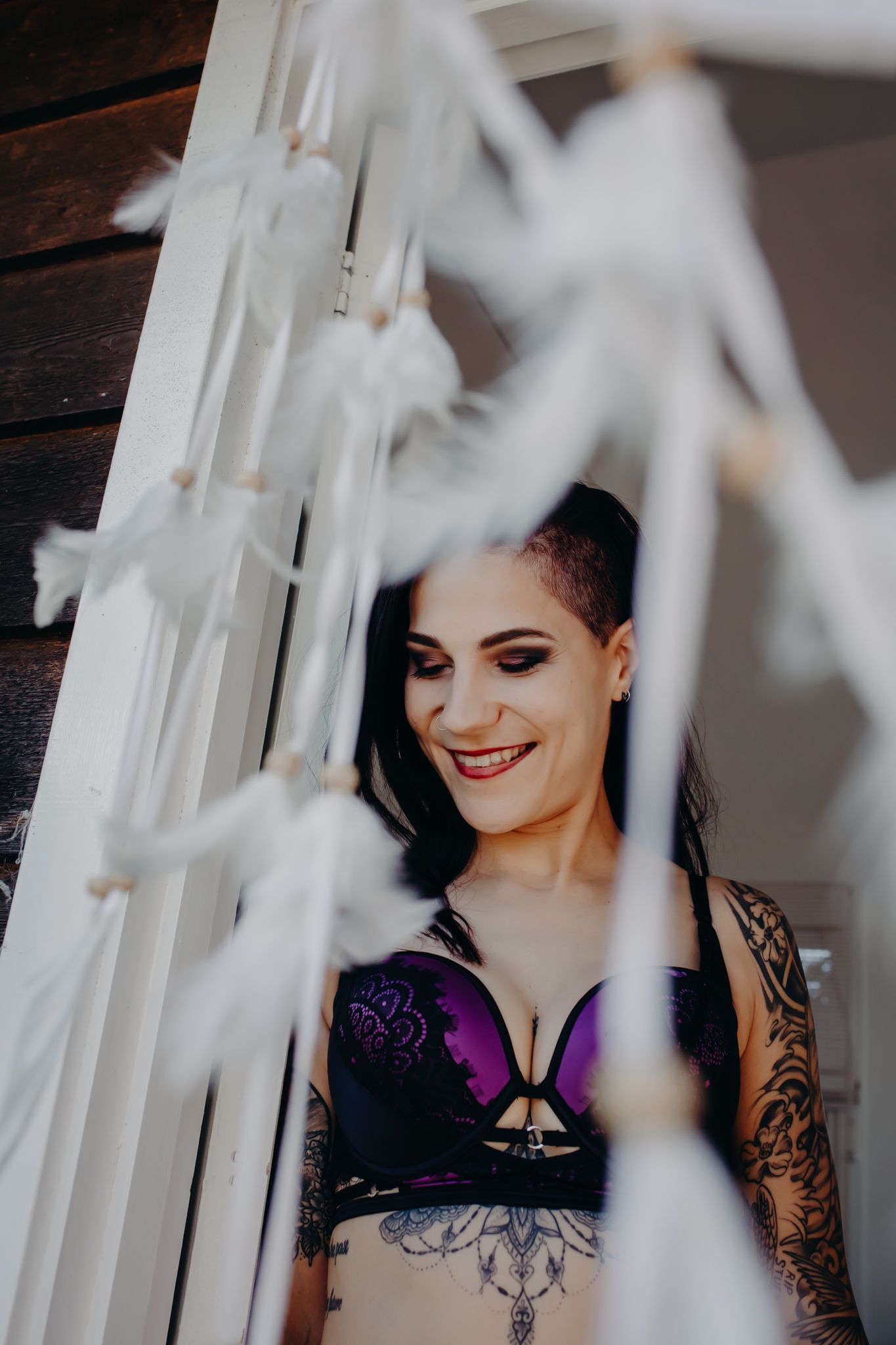 Artistic boudoir photo through white sheer curtains showing purple lingerie and tattoos.
