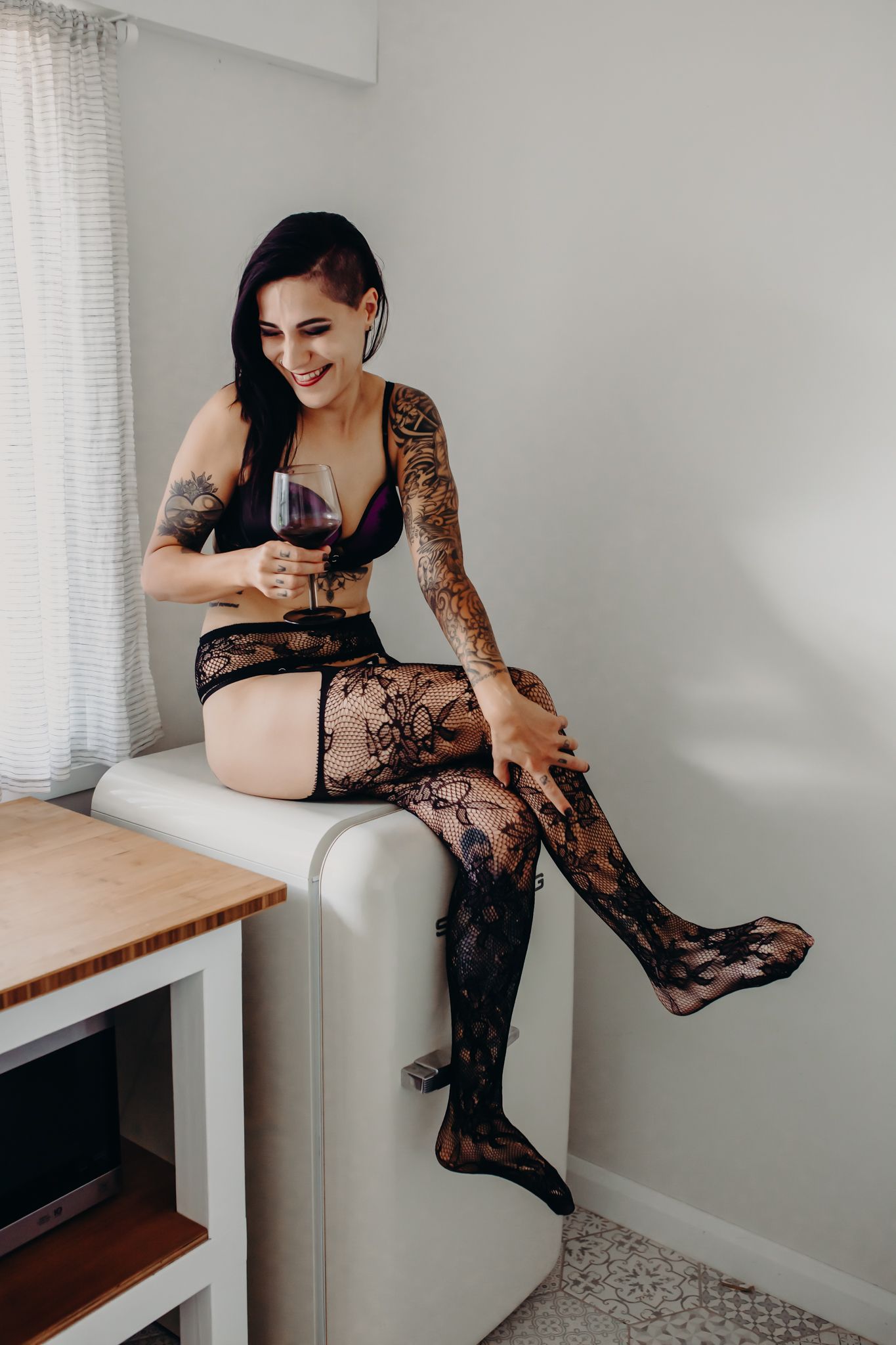 Woman in black lace lingerie sitting casually on white counter with wine glass.