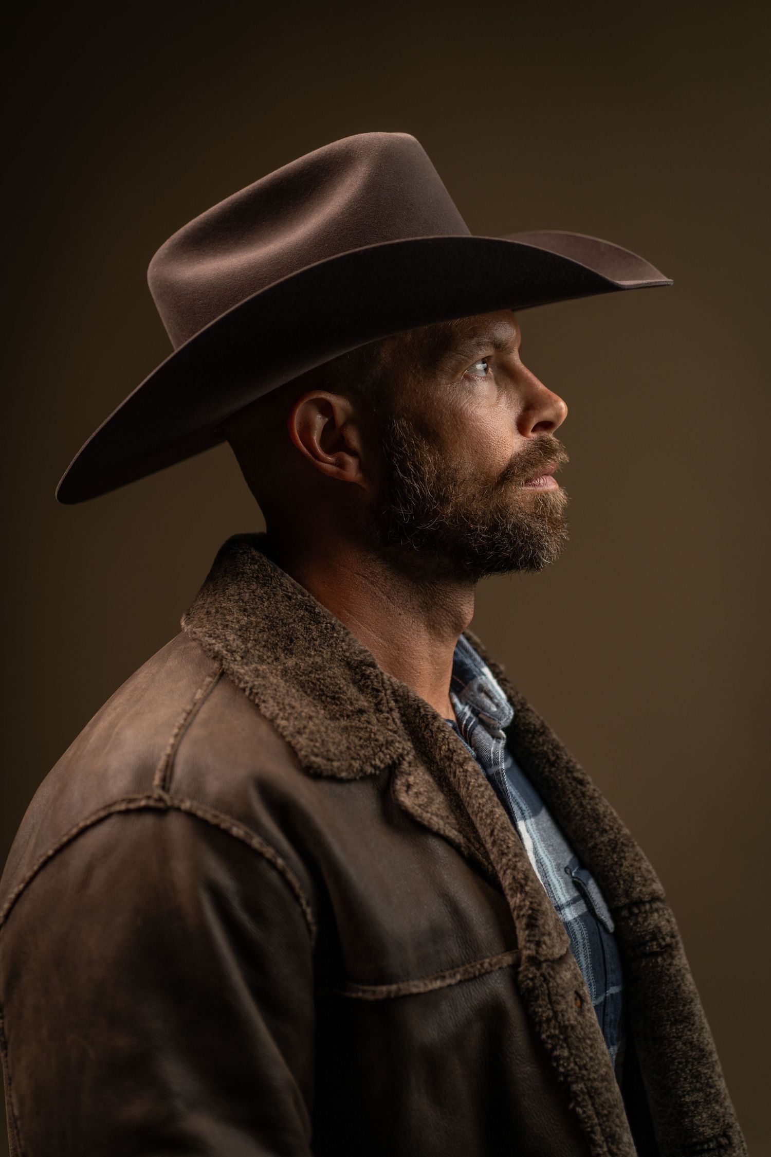 Side profile portrait of person in cowboy hat and shearling jacket against dark background.