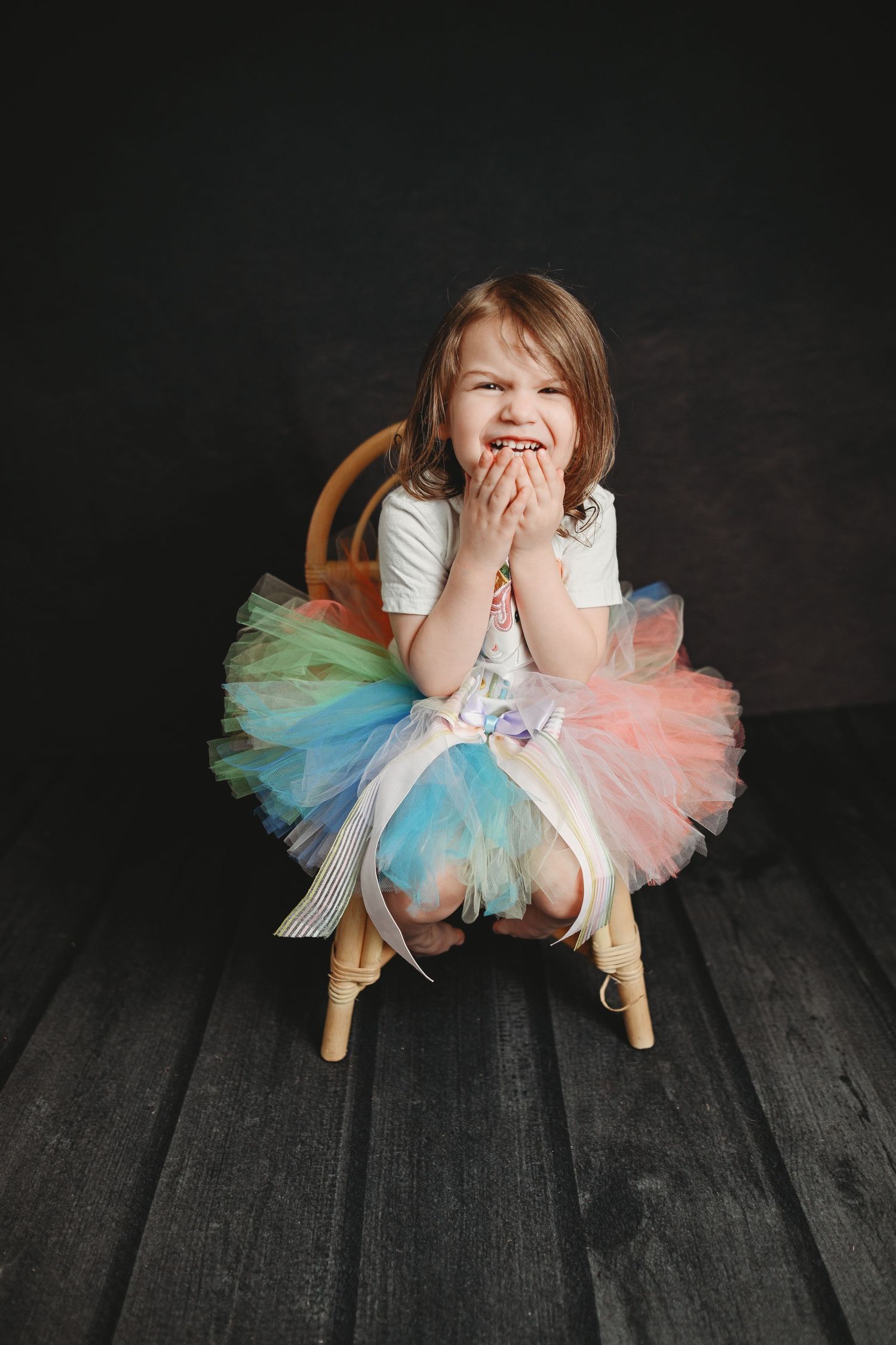 Child in colorful tutu dress poses playfully on wooden floor against dark background.