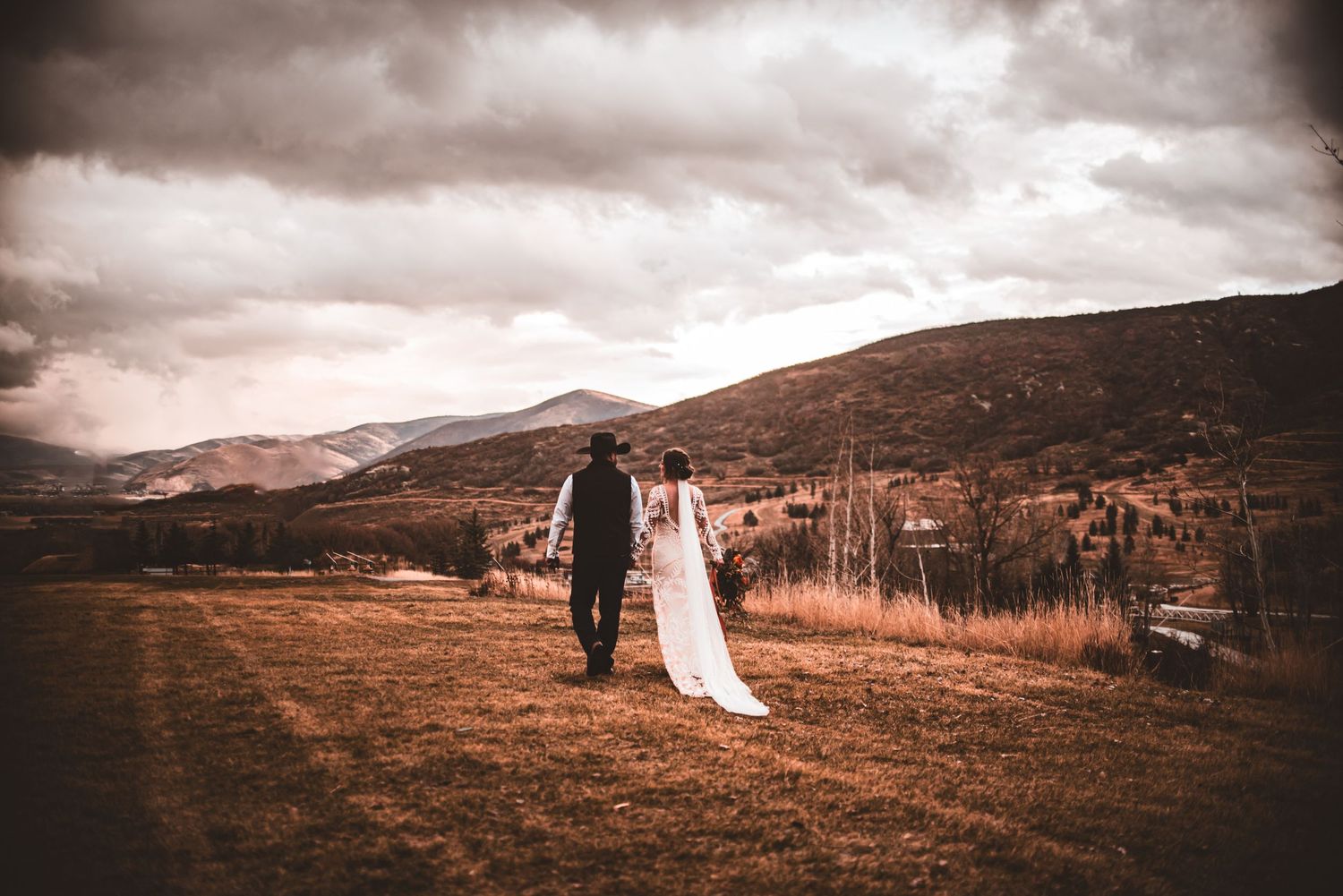 A newlywed couple walks hand in hand across a mountain meadow under moody skies.
