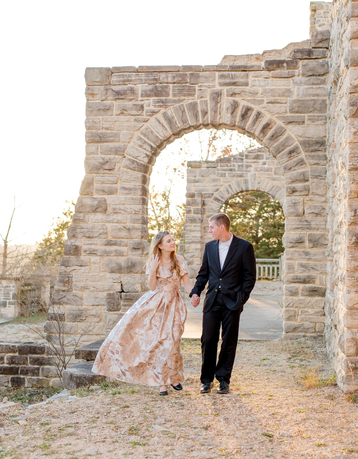 A couple in formal attire walks and laughs together beneath a rustic stone archway at sunset.