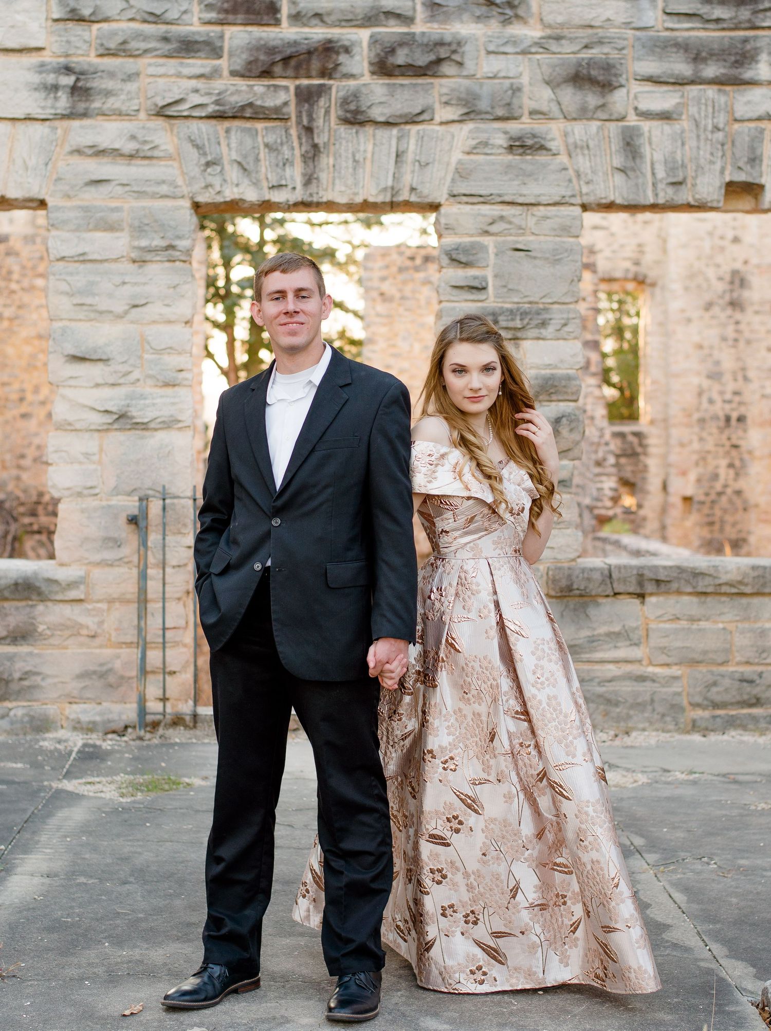 A couple poses at formal event wearing black suit and blush pink brocade gown against stone wall backdrop.