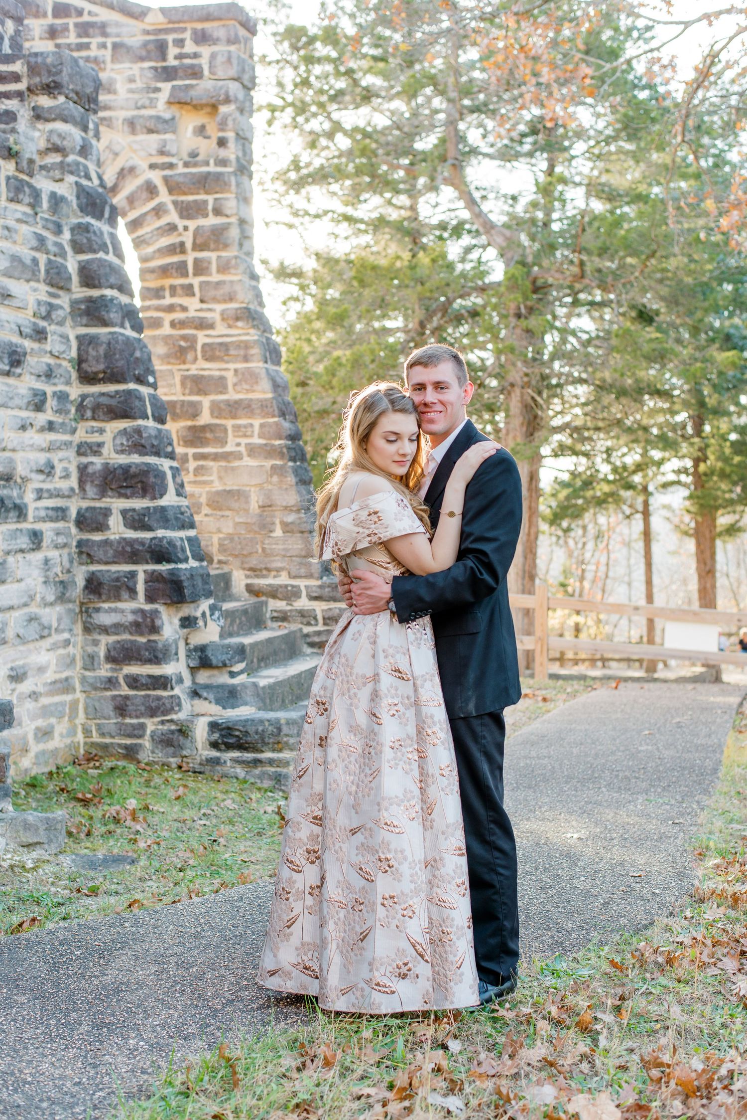 A couple in formal attire embraces near a stone wall and autumn trees along a path.