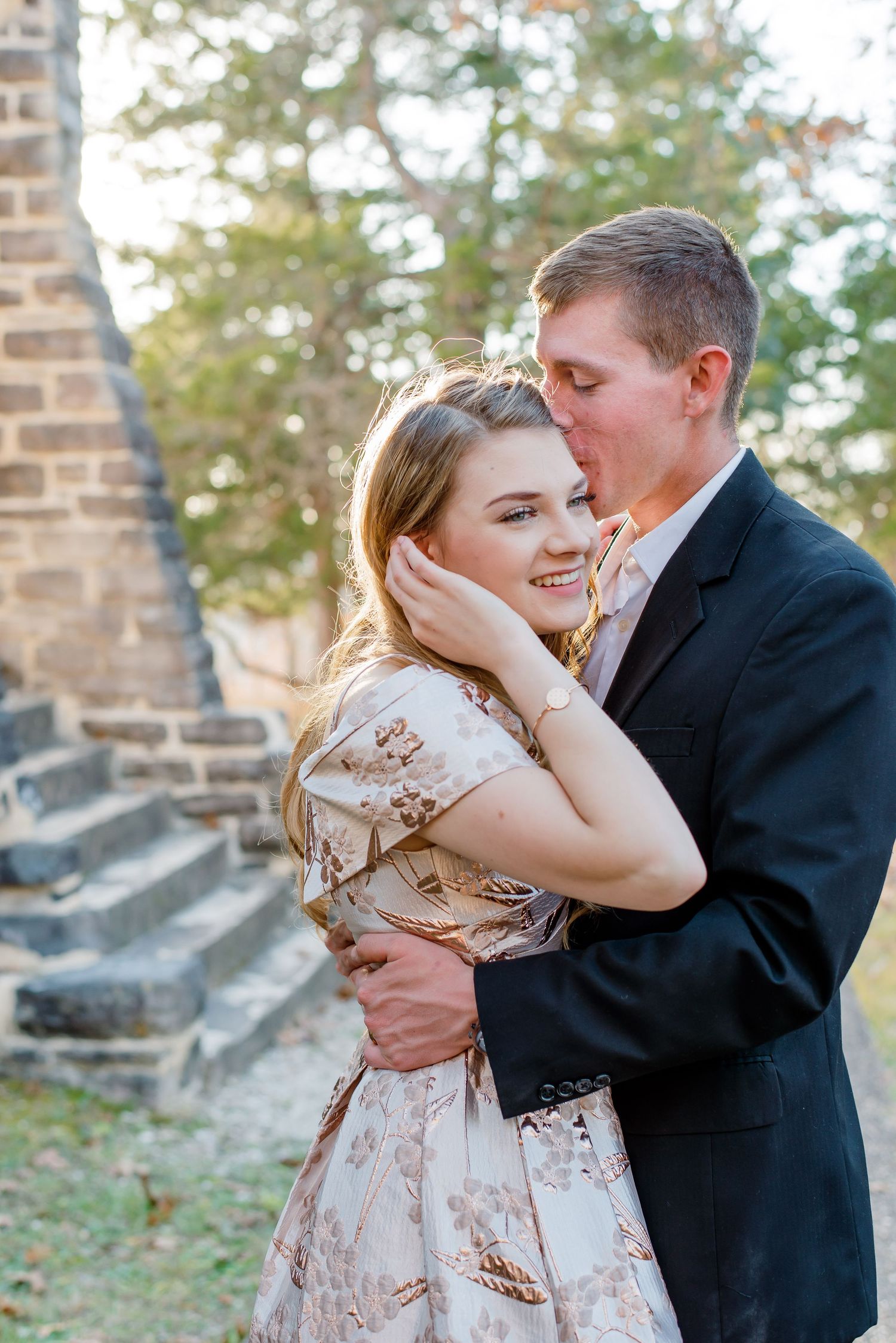A romantic couple shares an intimate moment on stone steps surrounded by greenery, with one wearing a patterned dress.