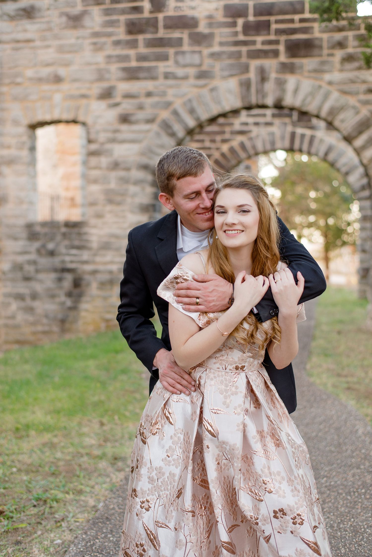 A couple in formal attire embraces in front of a stone archway on a path during their romantic photoshoot.