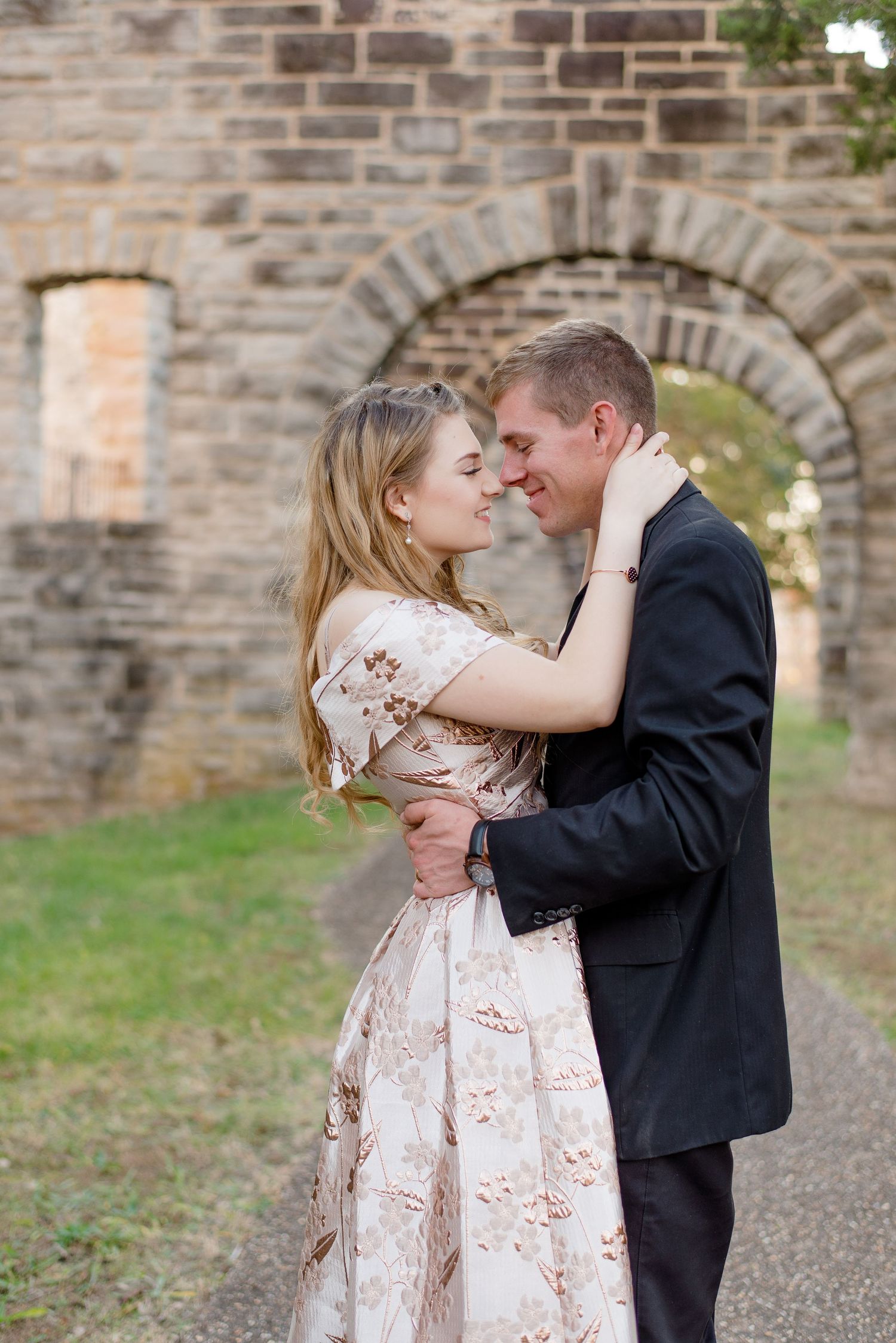 A romantic couple in formal attire embrace near a stone archway during an outdoor photoshoot.