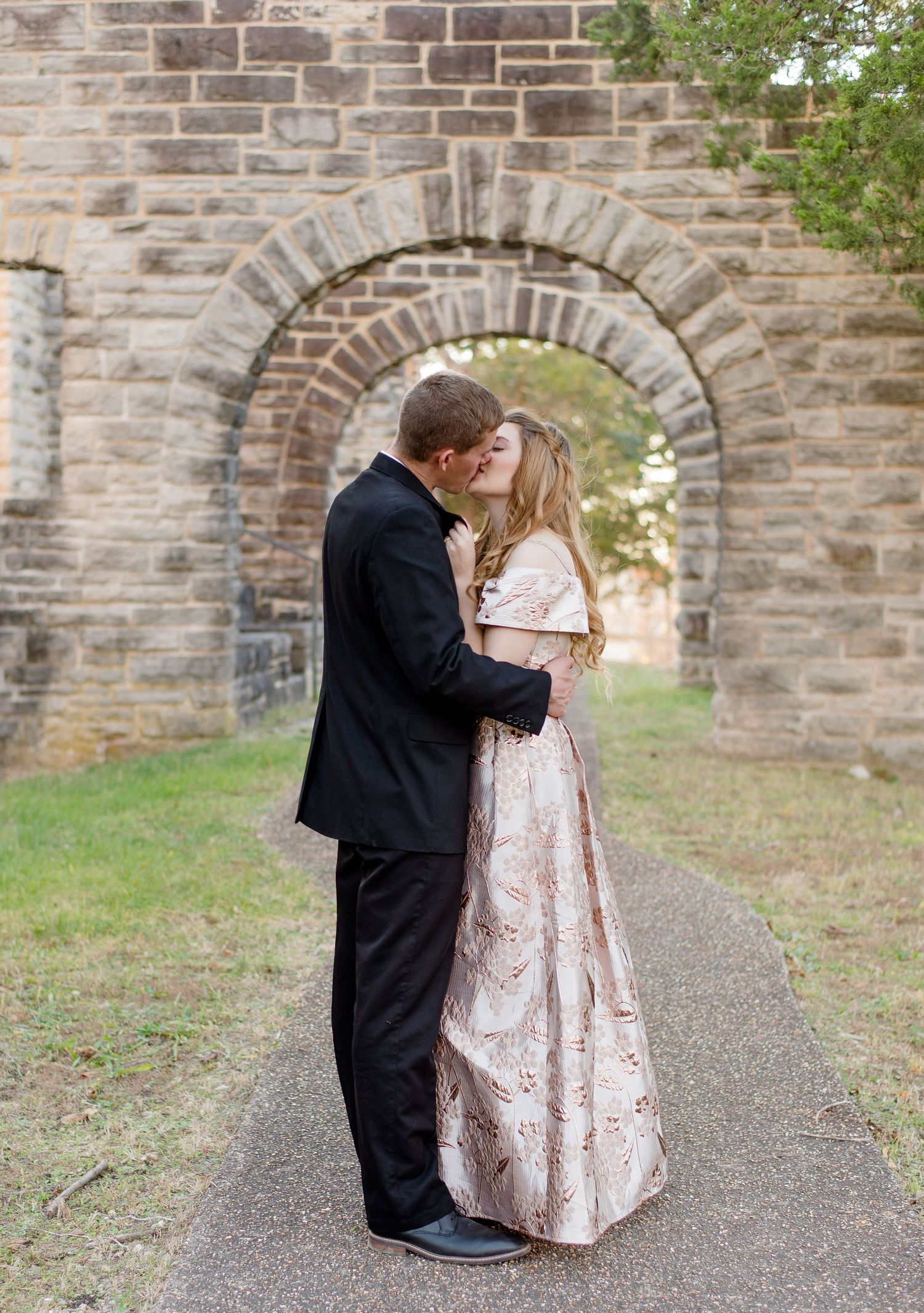 A romantic couple embraces and shares a kiss under a stone archway on a path during a formal outdoor photoshoot.