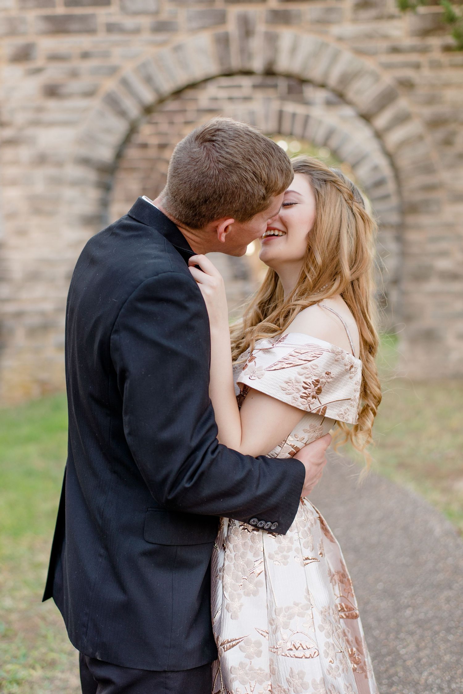 A romantic couple embraces and kisses under a stone archway, with the woman wearing a blush lace dress.