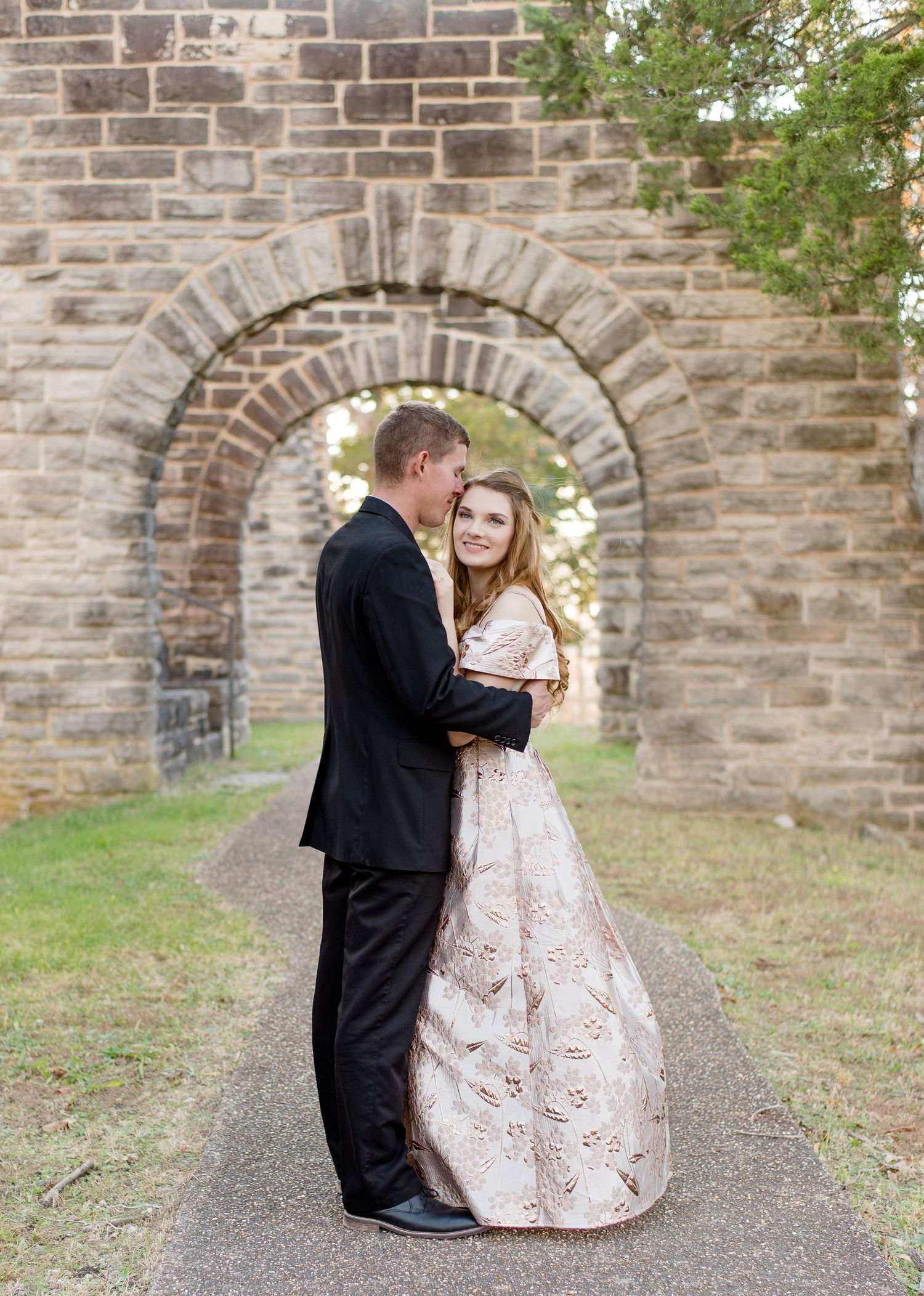 A romantic moment captured under a stone archway with a couple in formal attire on a paved path.