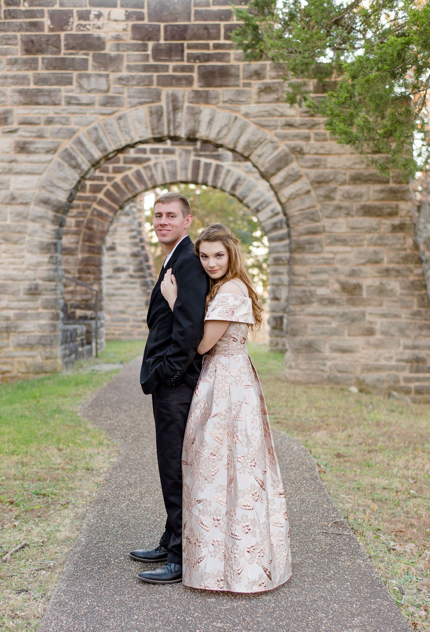 A couple in formal attire poses in front of a stone archway on a path, with the man in black and woman in pink gown.