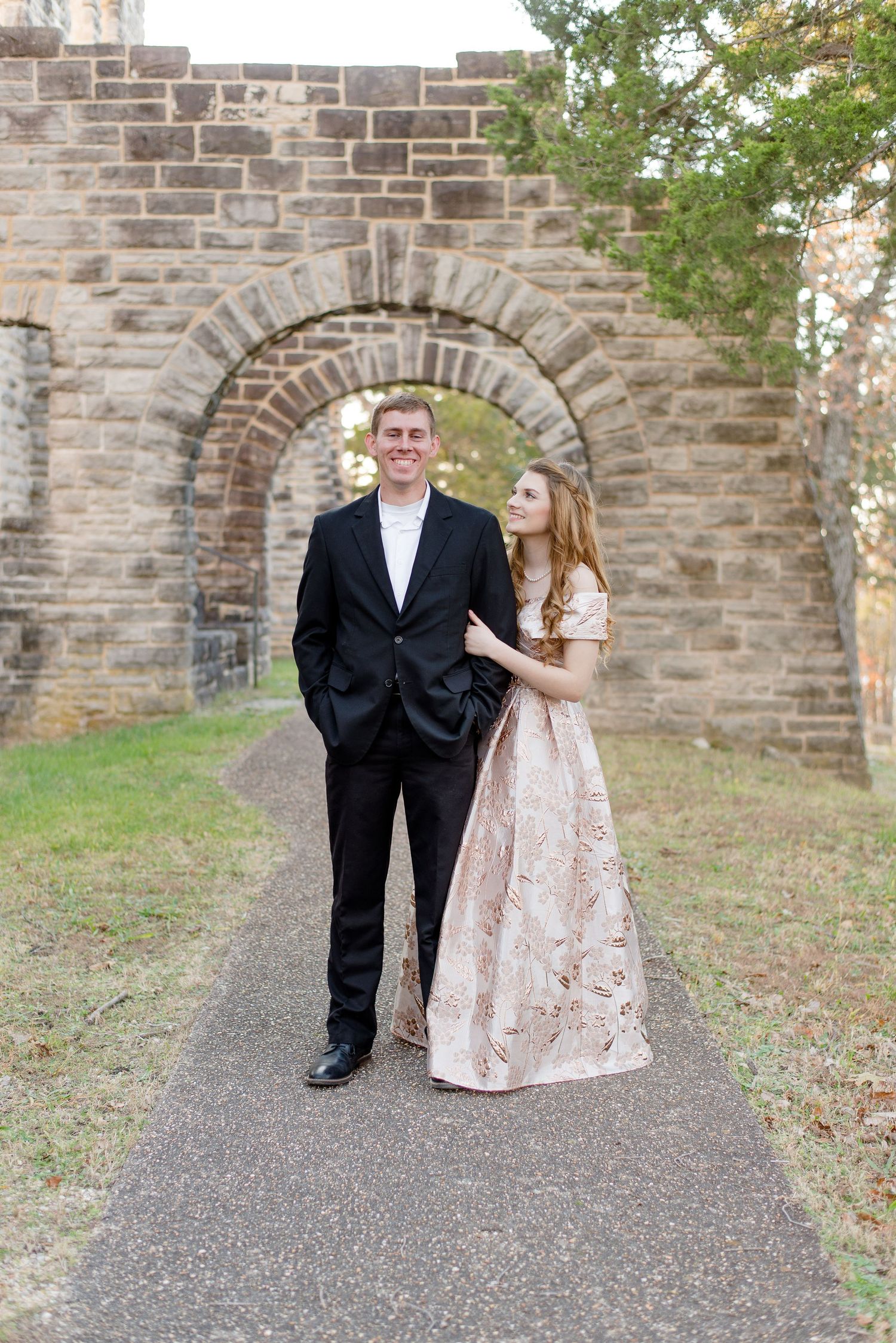 A couple in formal attire stands beneath a stone archway on a paved path during an outdoor photoshoot.