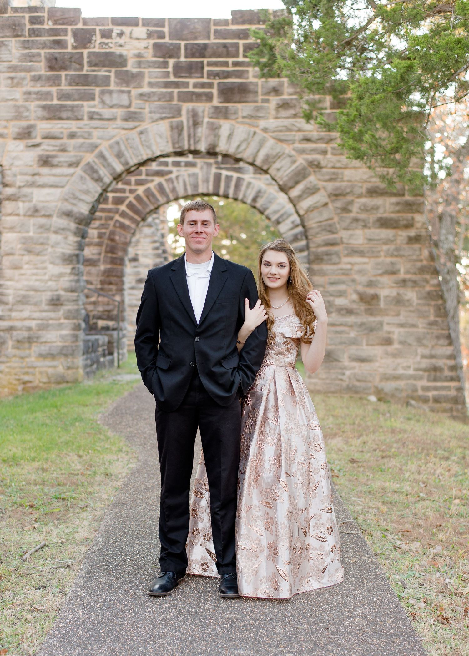 A formal couple poses under a stone archway on a paved path, wearing a black tuxedo and rose gold evening gown.