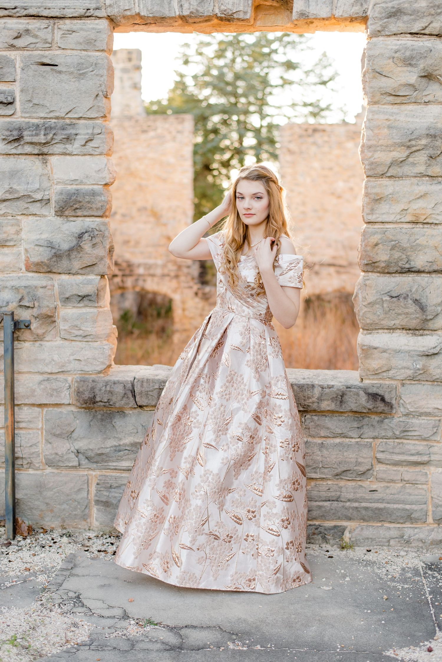 Elegant metallic rose gold ballgown with floral brocade pattern photographed in stone window archway at sunset.