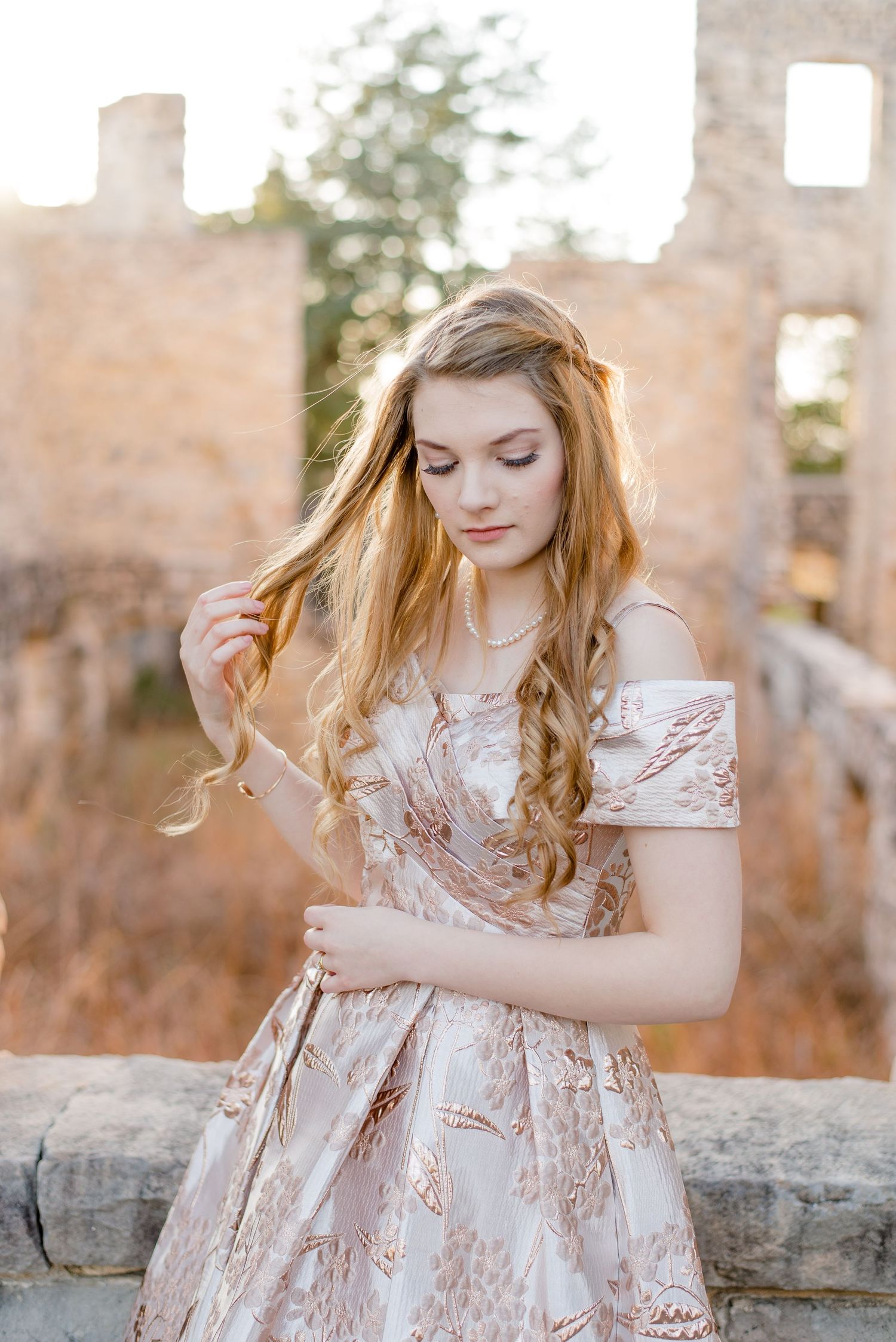 A rose gold brocade dress shimmers in golden sunlight against stone ruins with romantic vintage styling.