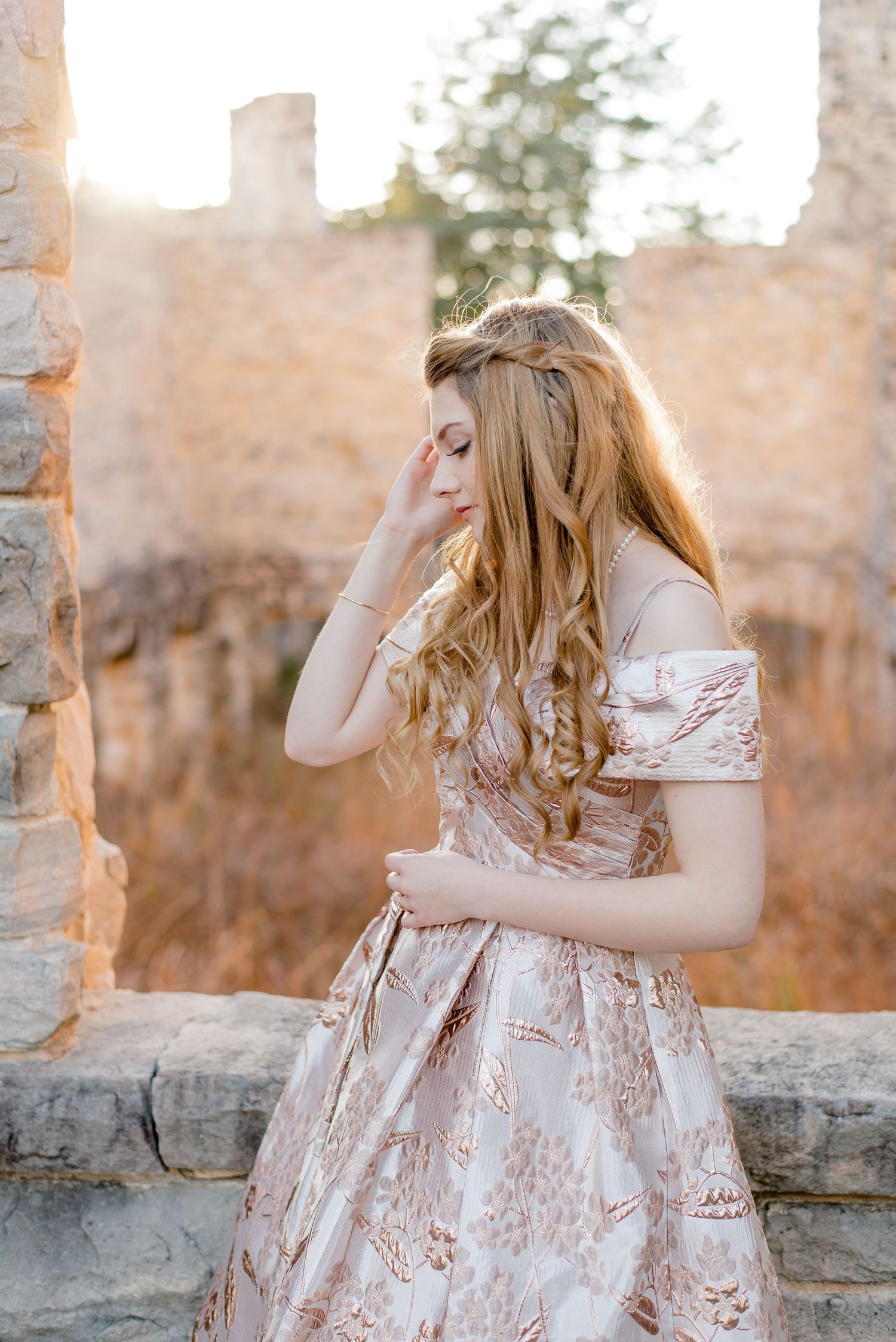Romantic portrait of someone wearing an ornate blush pink gown with lace details against stone ruins at sunset.