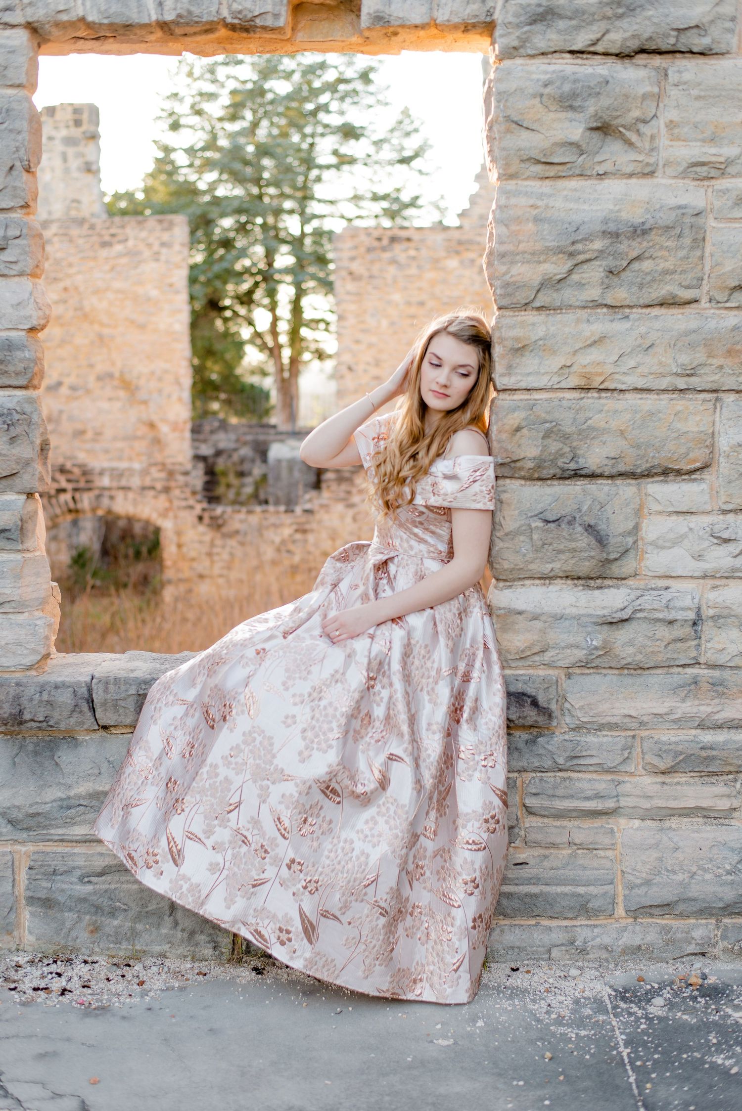 An elegant vintage-style gown with floral patterns is showcased against weathered stone ruins at sunset.