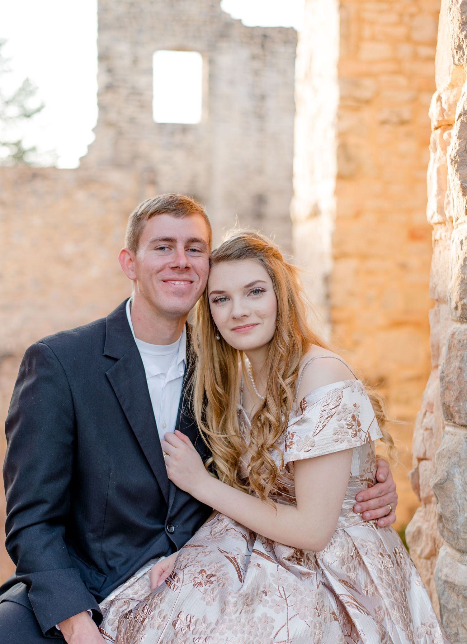A couple in formal attire pose together against stone ruins during a golden hour photo session.