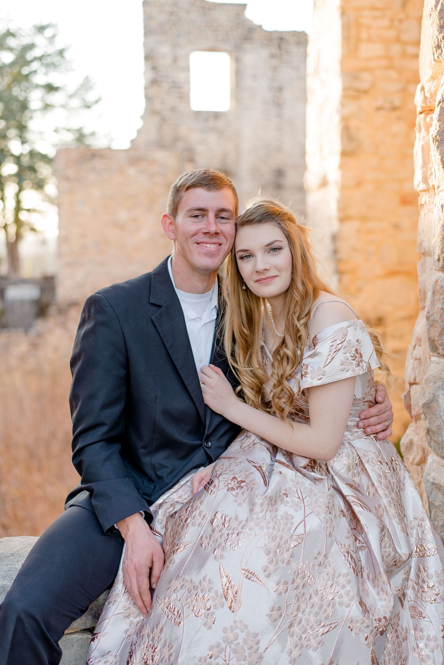 A couple sits together at a stone ruin during golden hour, dressed in formal attire with a vintage aesthetic.