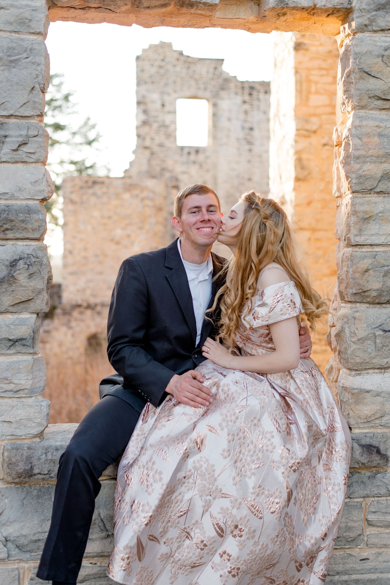 A romantic couple in formal attire share an intimate moment while sitting in a stone window frame of old ruins.