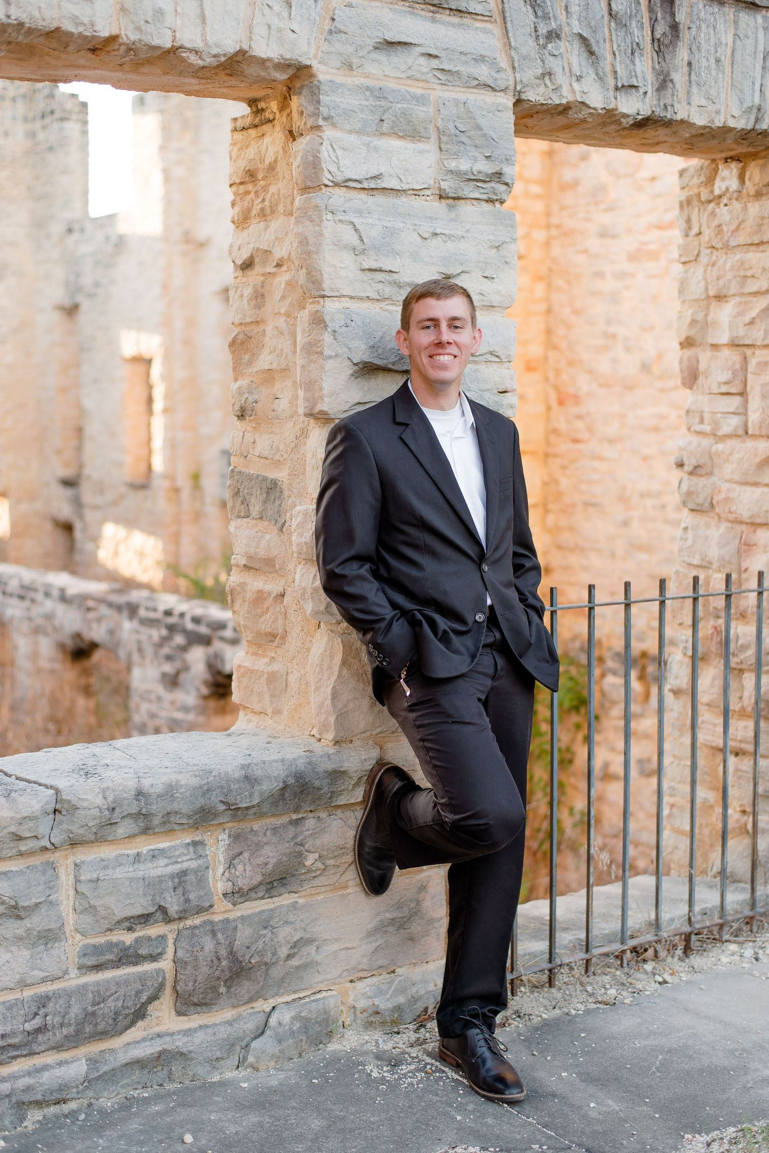 A person in a black blazer and dark pants poses casually against a stone wall with archways in an architectural setting.