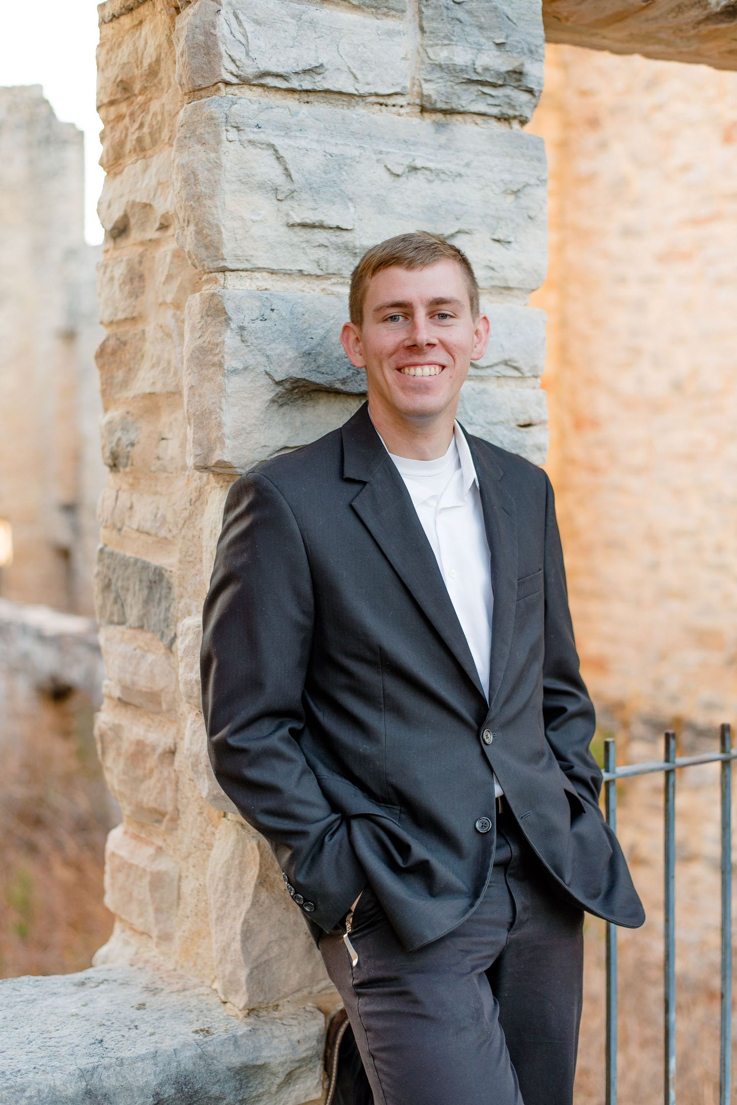 Professional portrait in a black suit and white shirt standing against a stone wall outdoors.
