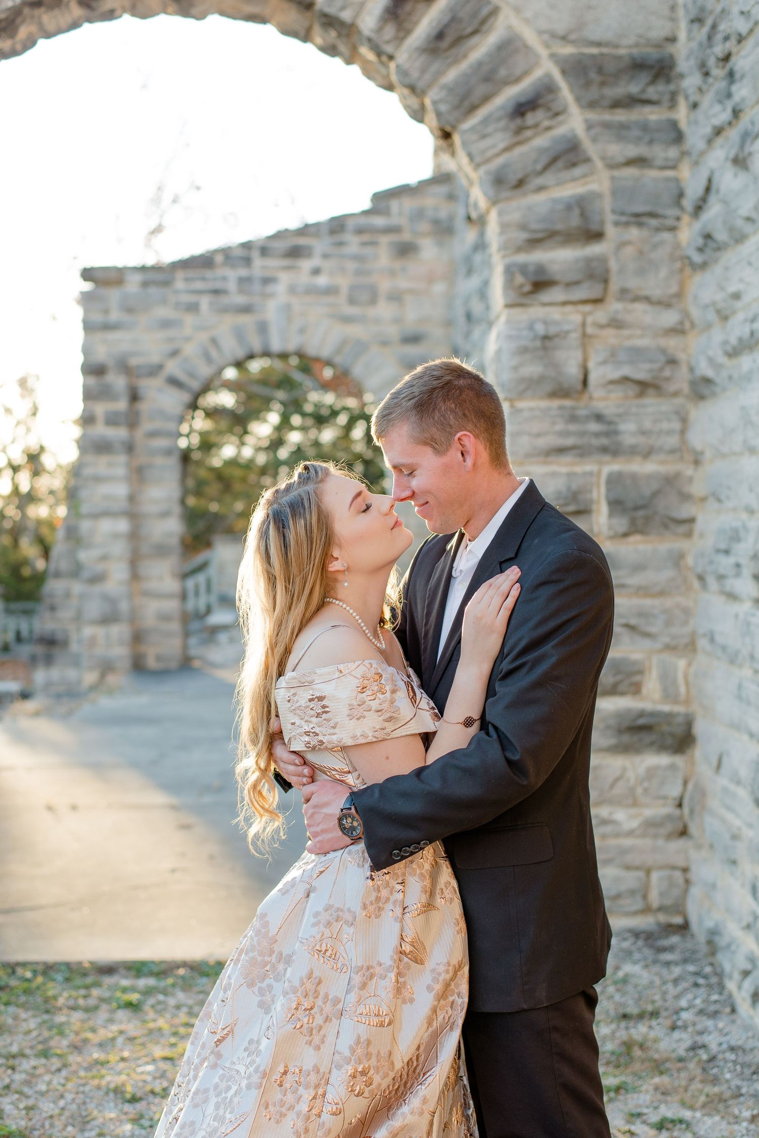 Romantic couple embraces under stone archway at sunset wearing formal attire and vintage-style dress.