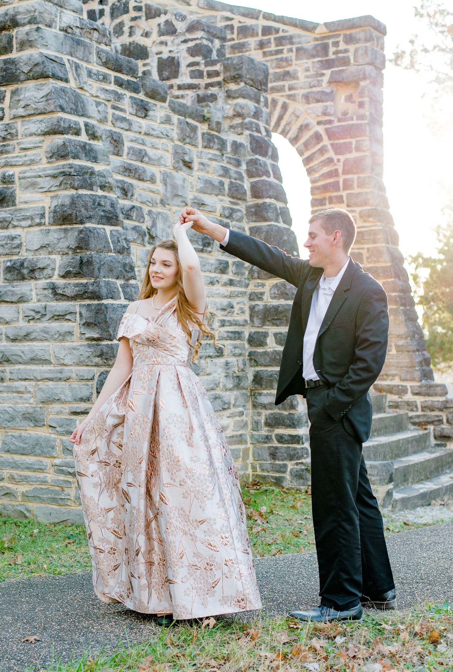 A couple in formal attire poses by a stone wall, with the woman in a blush ball gown and the man in a black tuxedo.