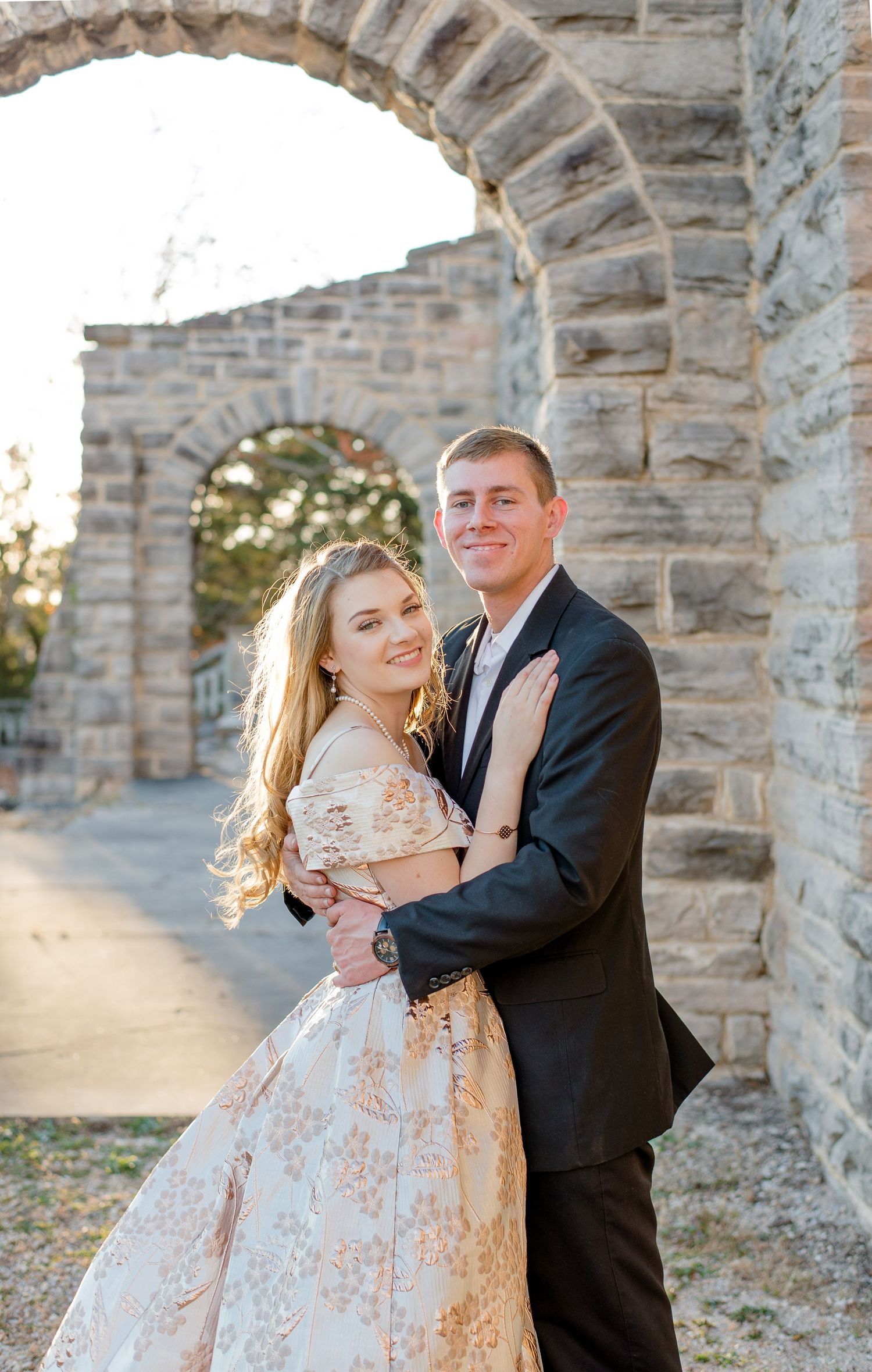 Couple embraces under stone archway at sunset, bride wearing lace floral gown and groom in classic black suit.