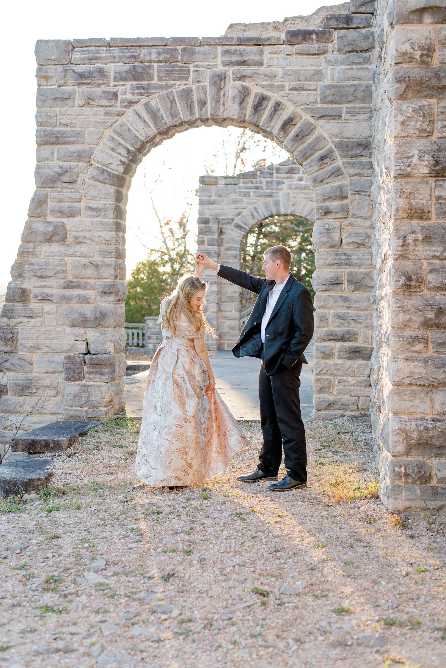 A couple in formal attire stands beneath a stone archway during sunset, dressed in a gold gown and black suit.
